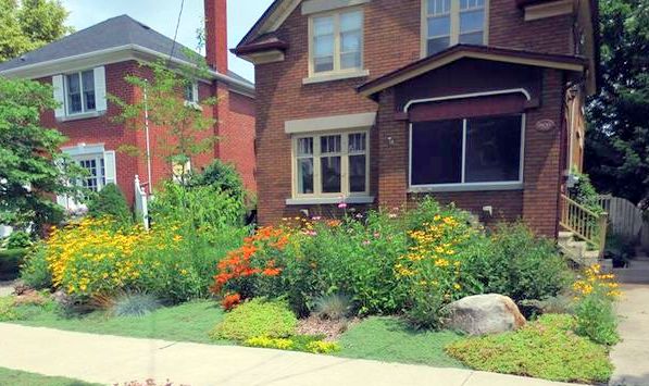 A brick house with a lush garden full of orange and yellow flowers. A sidewalk runs in front, and there's a clear sky overhead.