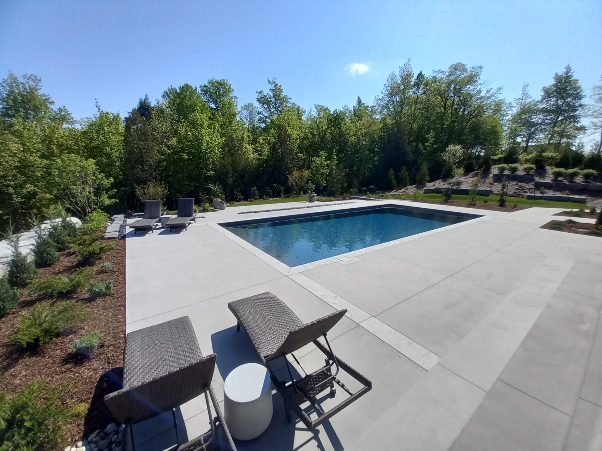 This image shows a modern backyard with a rectangular swimming pool, patio with lounge chairs, landscaped garden, and clear blue skies.