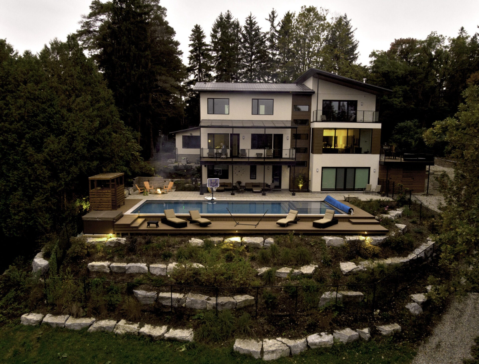 A luxurious multi-story house with large windows, surrounded by trees, featuring a pool, outdoor furniture, and terraced gardens in a tranquil setting.