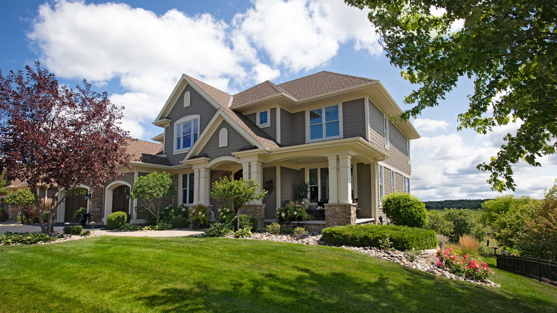 A large two-story house with a porch, gabled roofs, and stone accents surrounded by manicured lawn, landscaping, and a clear blue sky.