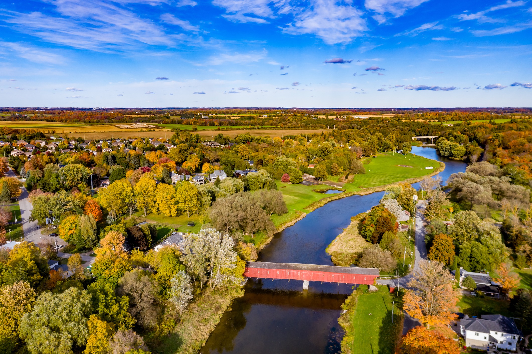 Aerial view of a landscape during autumn with a red covered bridge over a river, colorful trees, houses, and expansive fields under a blue sky.