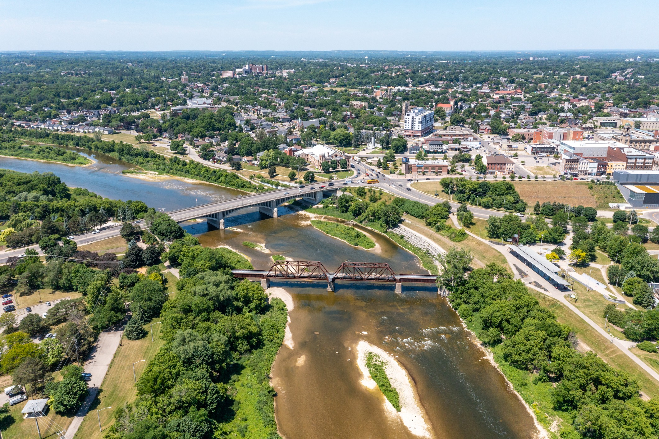 An aerial view of a sunny town with two bridges over a river, one operational and the other appears old and disused, surrounded by greenery and buildings.