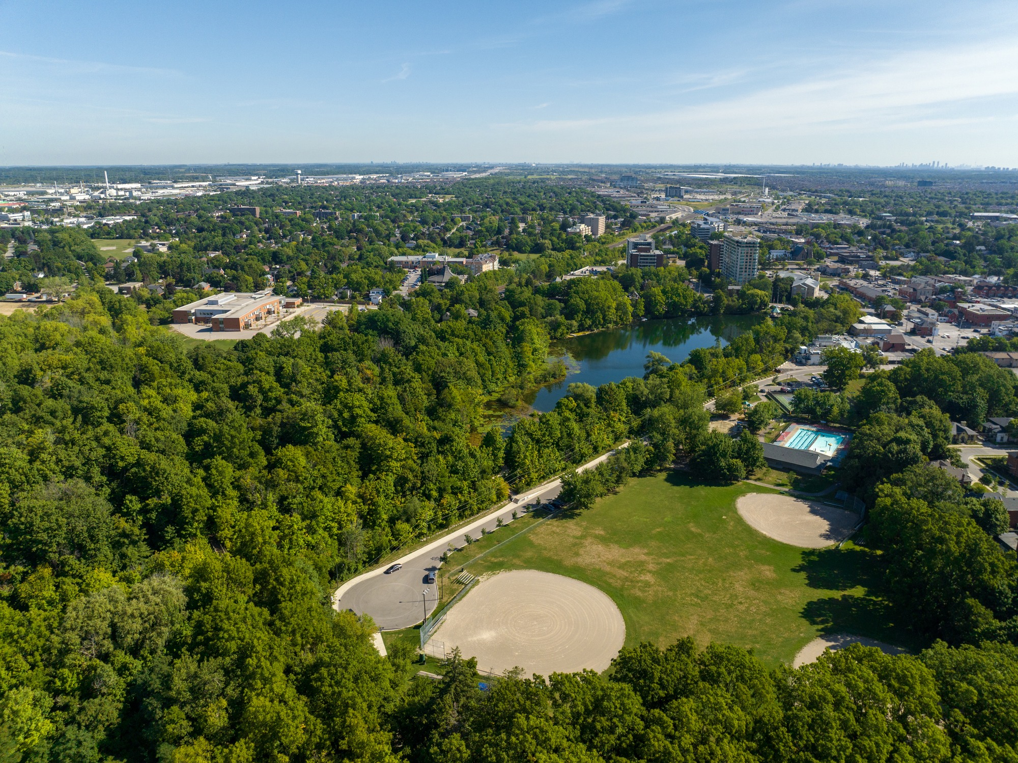 Aerial view of a lush, green urban park with a lake, buildings, swimming pool, and a clear blue sky, showcasing a vibrant community amidst nature.