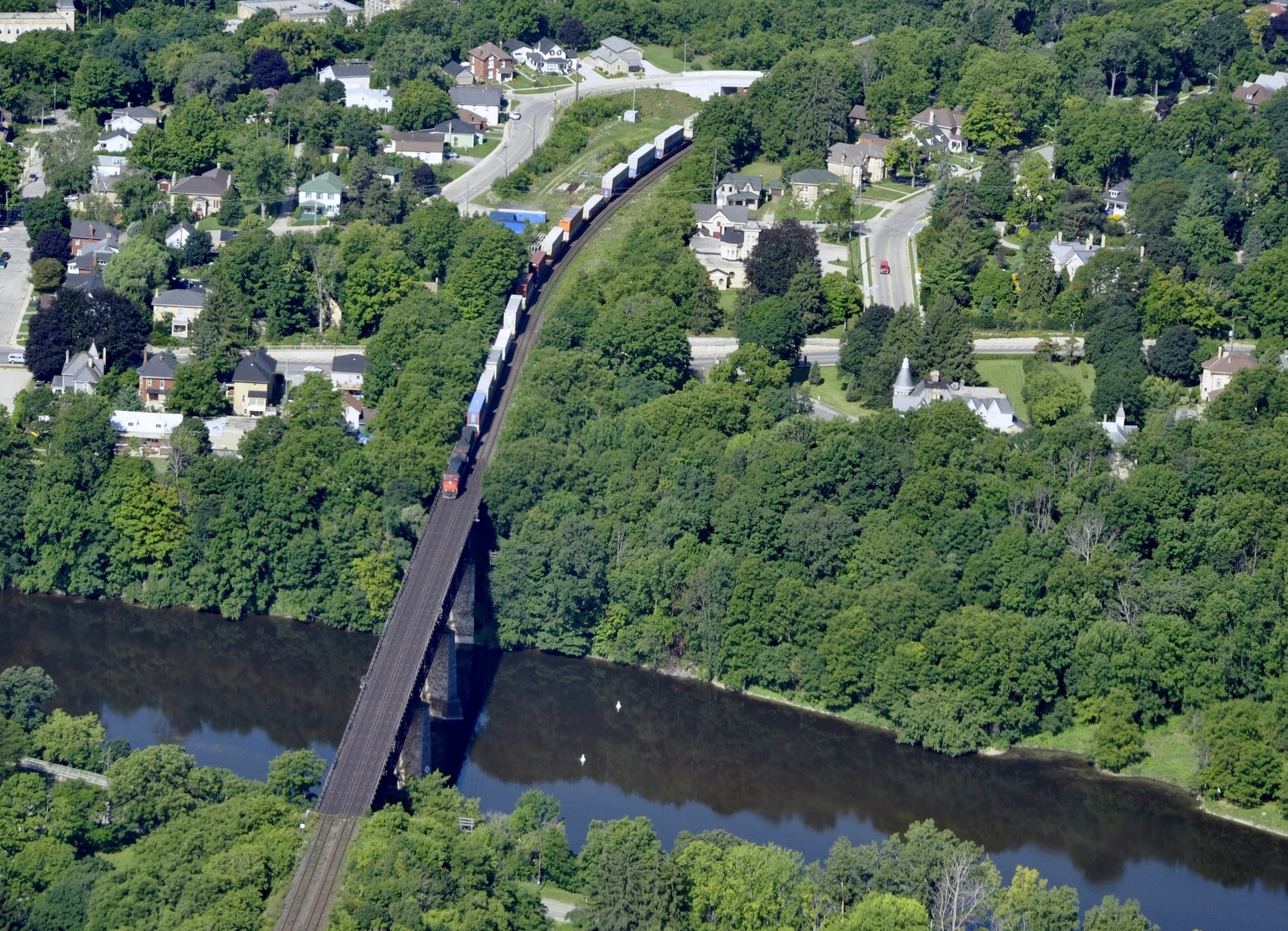 An aerial view of a cargo train crossing a bridge over a river, surrounded by lush greenery and residential houses in a tranquil neighborhood.