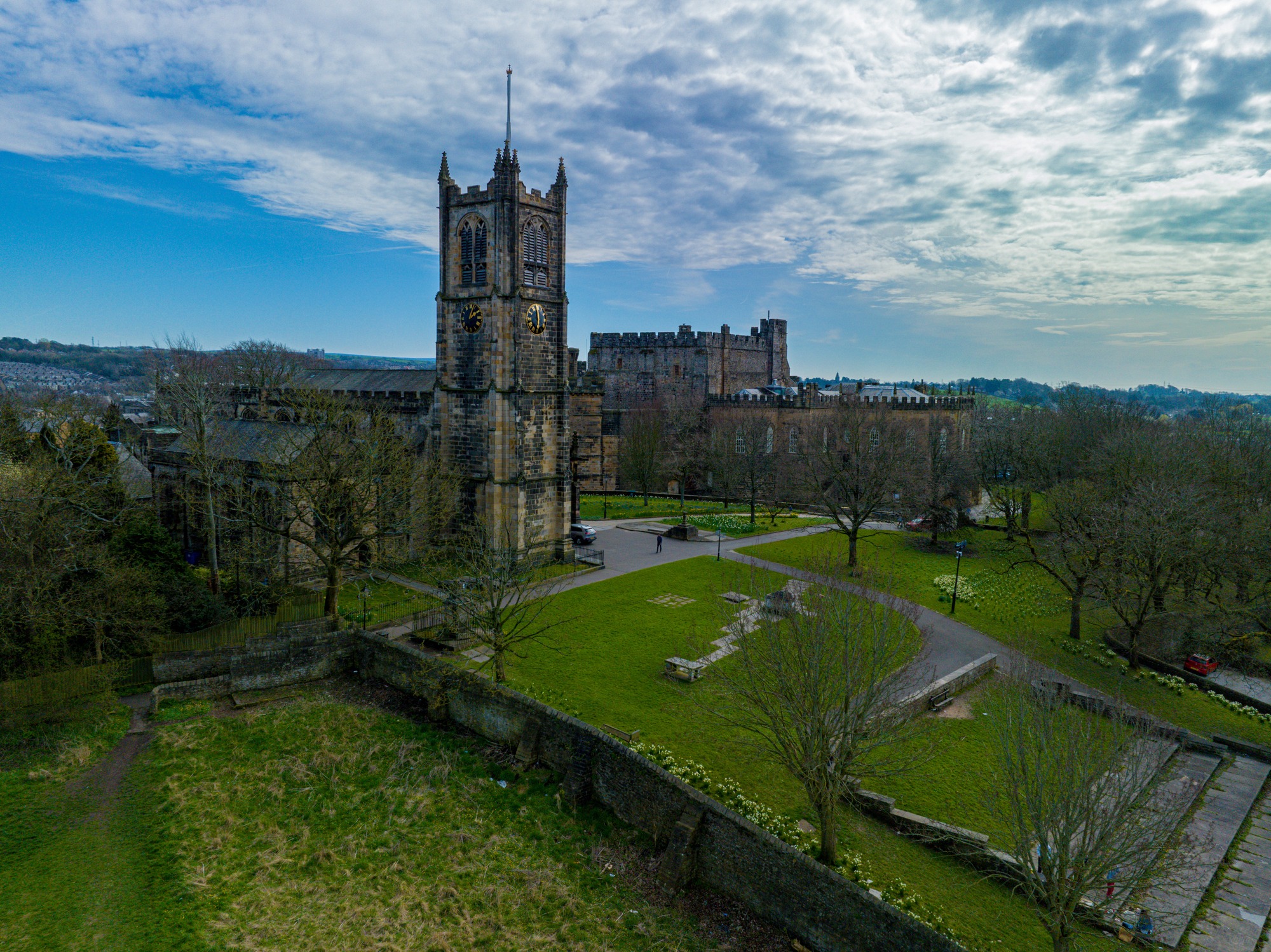 Aerial view of a historic cathedral and castle surrounded by trees, lawns, and walkways, with a flag atop, under a cloud-strewn blue sky.