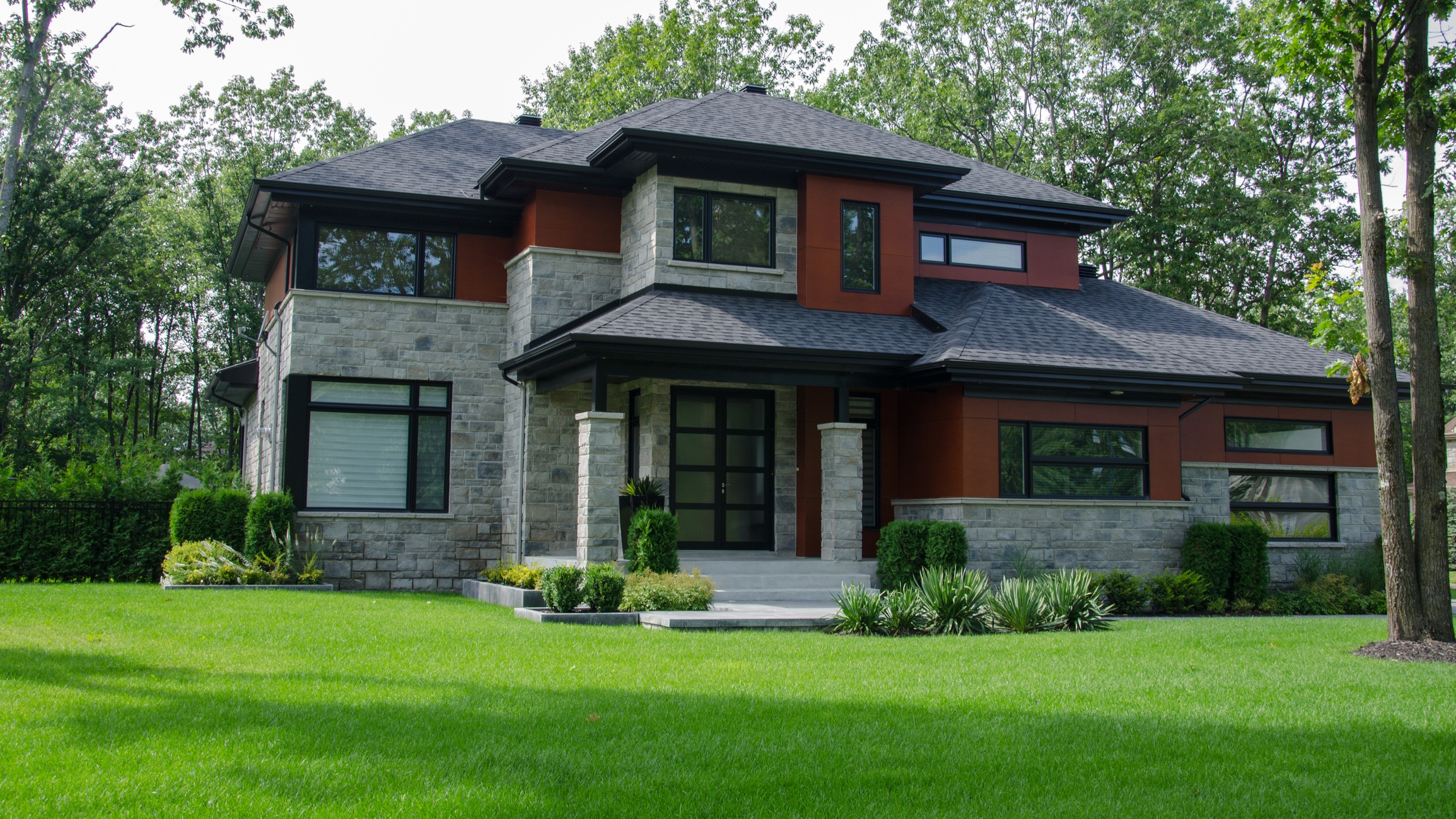 A modern two-story house with a dark roof, stone and red siding, large windows, surrounded by lush green lawn and ornamental plants.