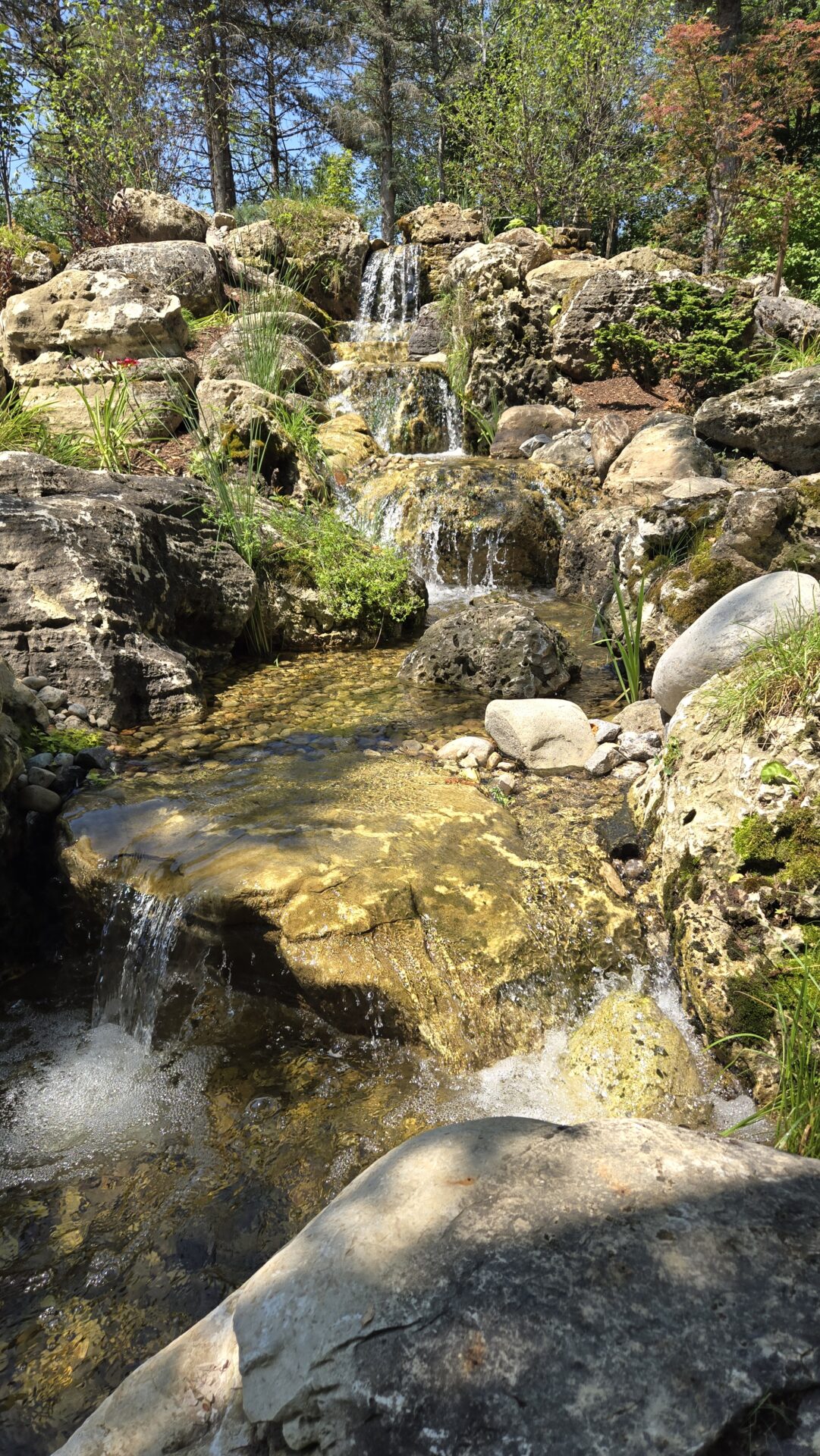 This image captures a serene man-made waterfall cascading over rocks surrounded by greenery under a bright blue sky on a sunny day.