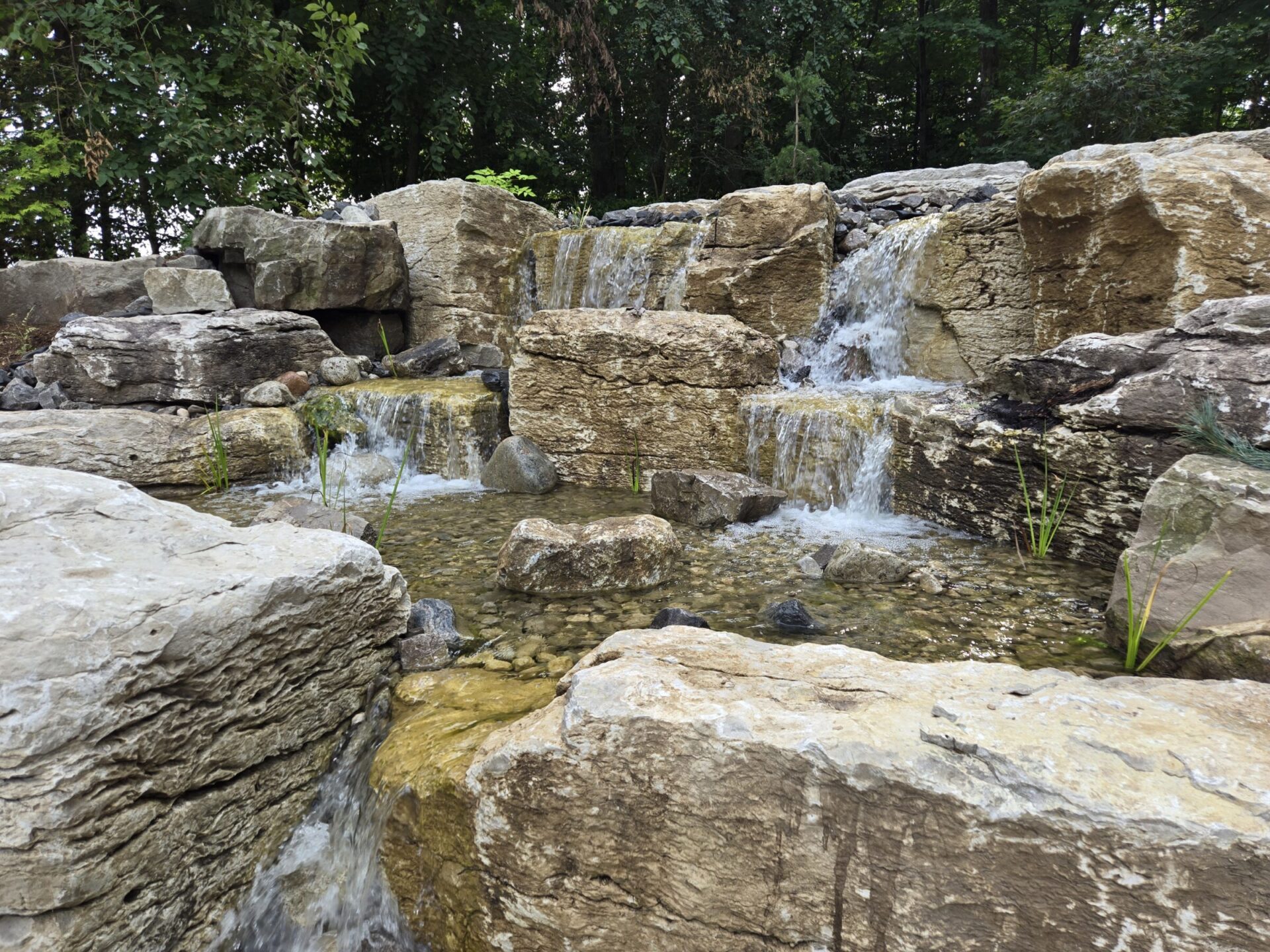 A cascading artificial waterfall with water flowing over layered limestone rocks, surrounded by greenery, in a serene outdoor setting.