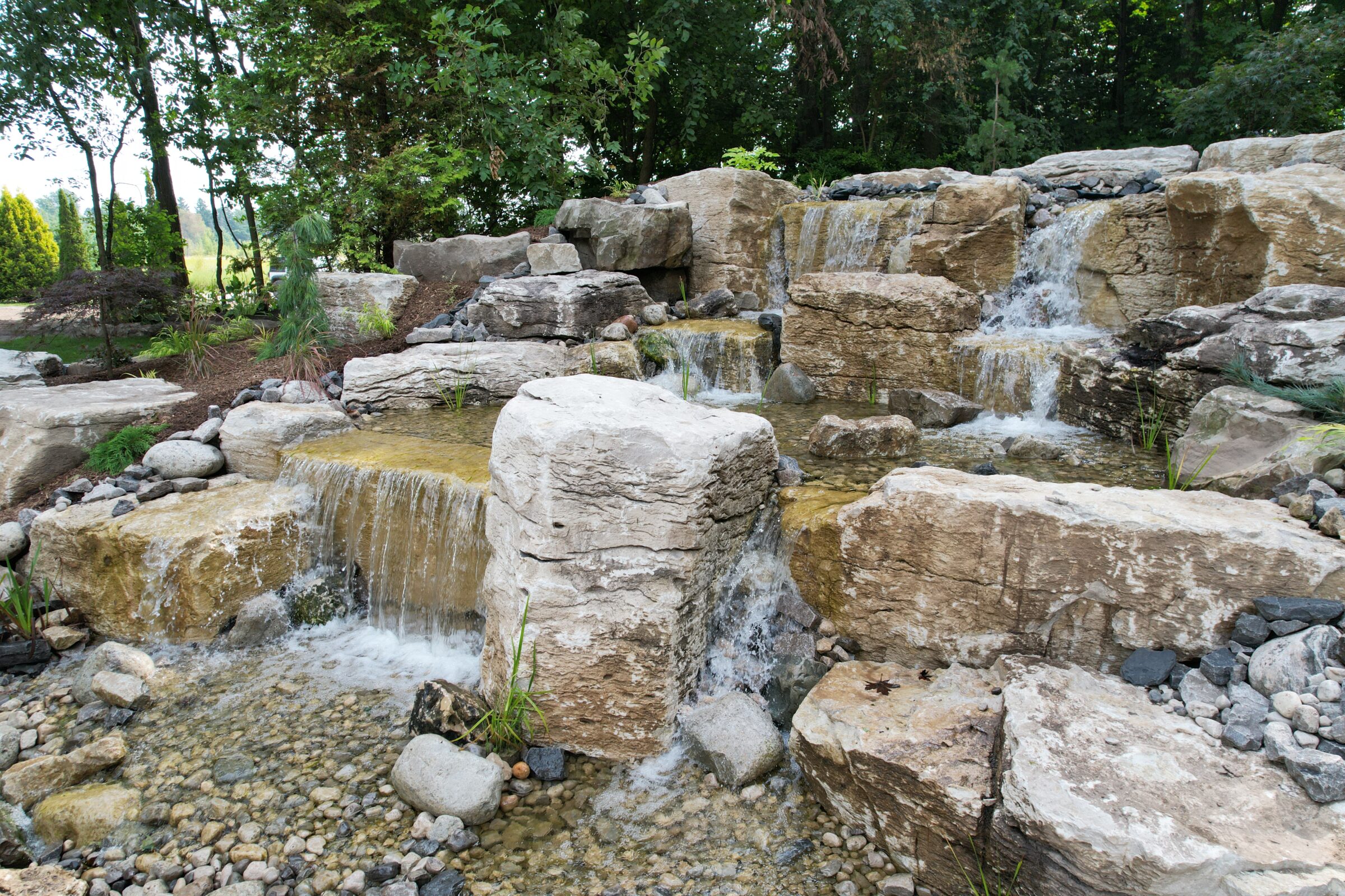 A man-made waterfall with cascading streams over rugged rocks surrounded by greenery, creating a serene landscape in a garden setting.