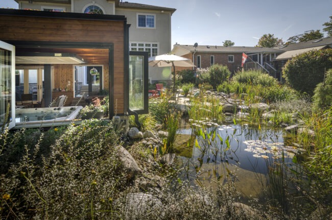 A peaceful backyard with a natural pond, lush vegetation, and a modern extension with large glass doors. Comfortable outdoor seating under an umbrella is visible.