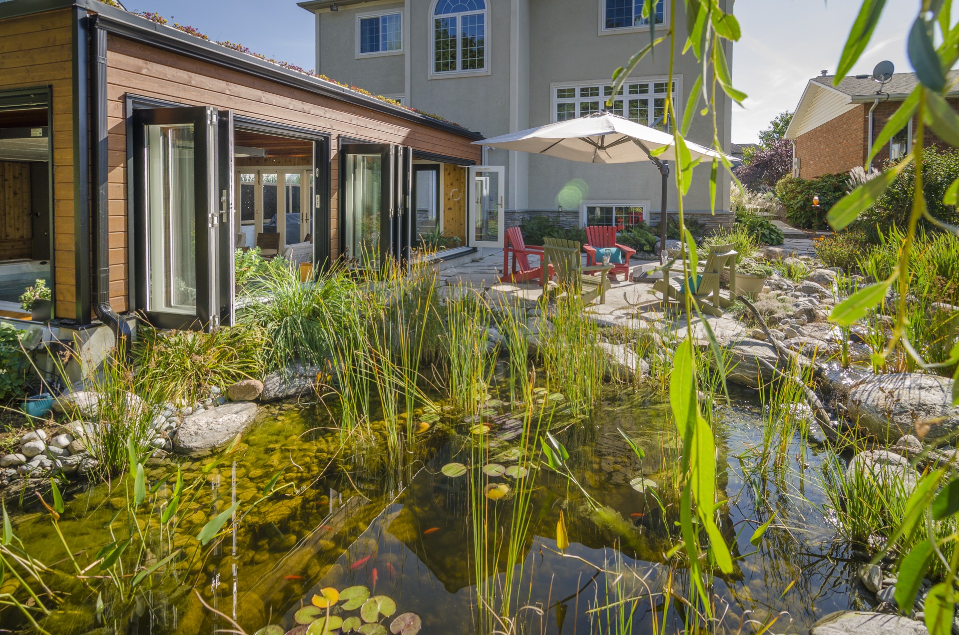 A serene backyard with a natural pond, surrounded by greenery, adjacent to a modern house with large windows, and a cozy patio area with red chairs.