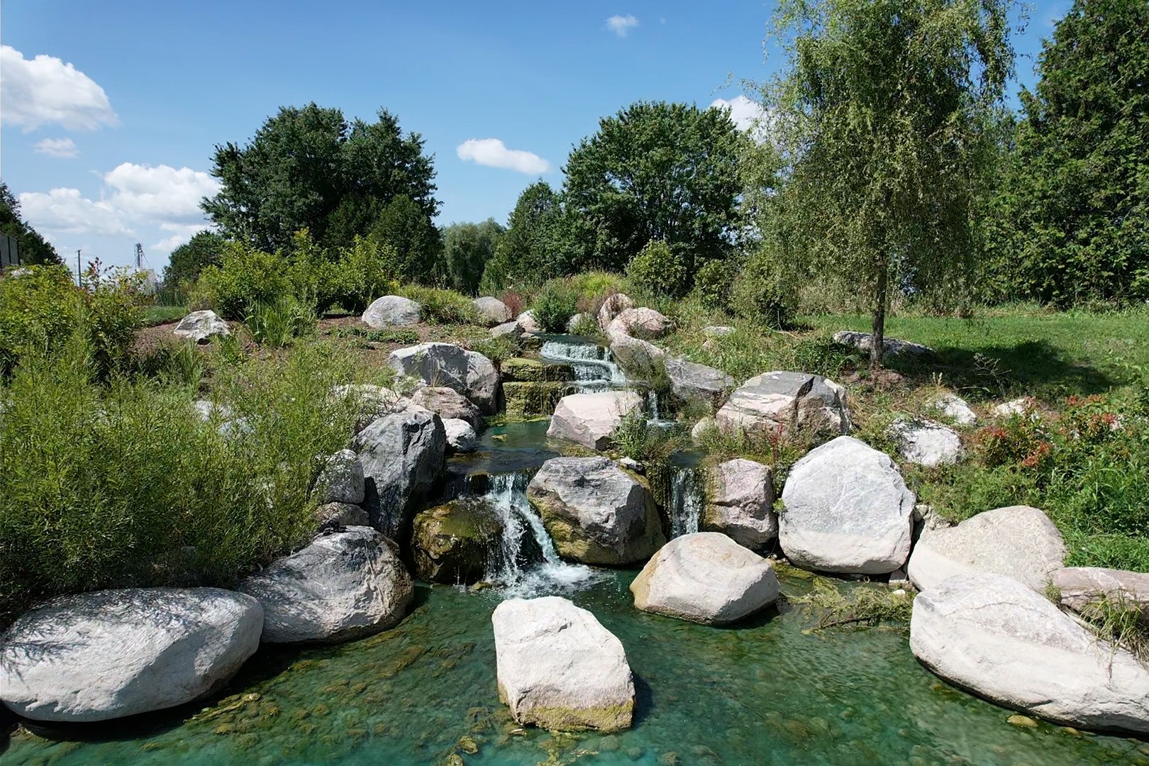 This image shows a tranquil rock-lined stream with clear water cascading over boulders, surrounded by green shrubbery under a blue sky with clouds.