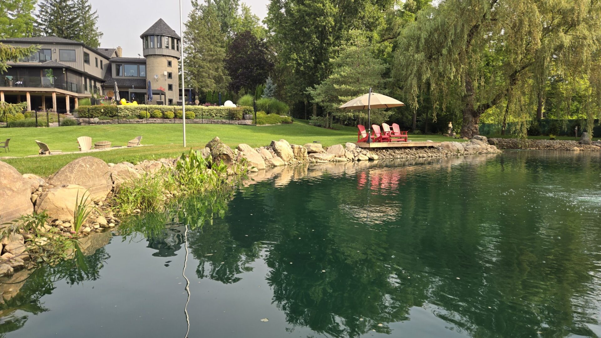 The image shows a tranquil pond with clear water, red chairs under an umbrella, a lush lawn, a stately home with a turret, and mature trees.