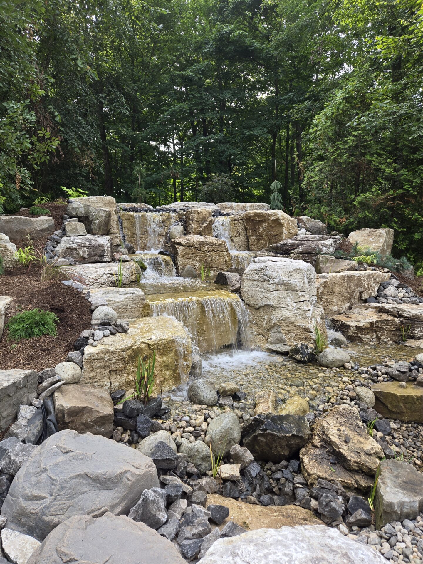 A serene waterfall cascades over large rocks, surrounded by lush greenery and forested trees, creating a tranquil natural landscape.