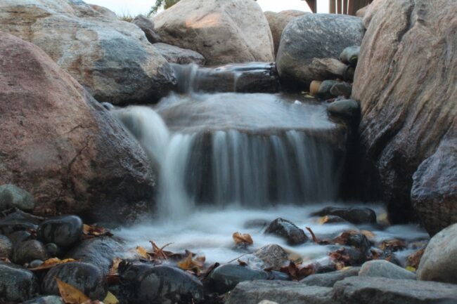 A small waterfall cascades over rocks, surrounded by smooth stones and dry leaves, creating a serene natural scene.