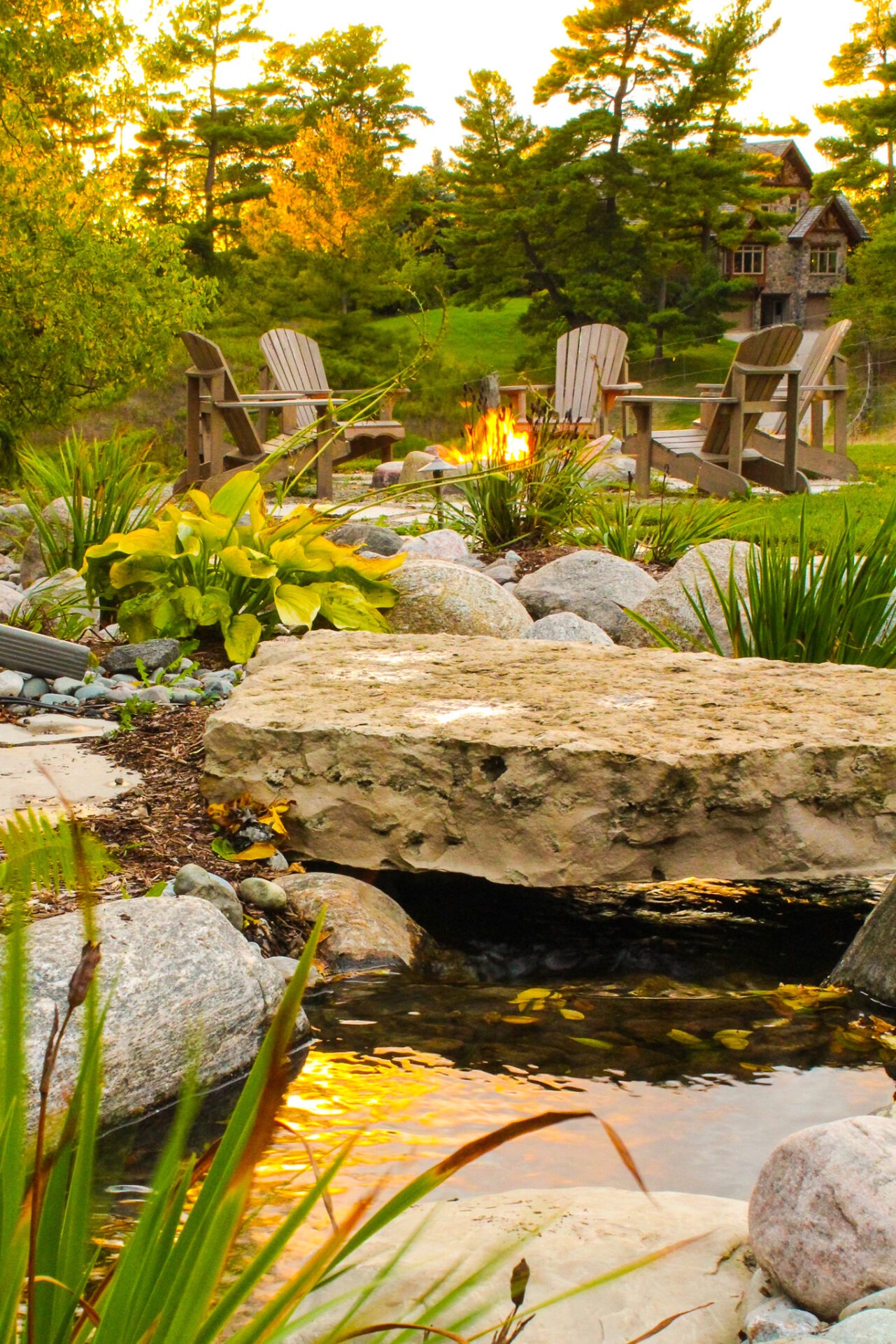 Four wooden chairs around a fire pit, surrounded by lush greenery and rocks, with a rustic cabin in the background during sunset.