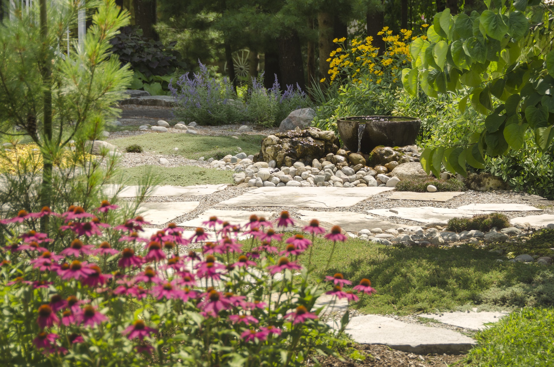 A serene garden with colorful flowers, stone pathways, and a small fountain, surrounded by lush greenery and vibrant blooms under sunny skies.