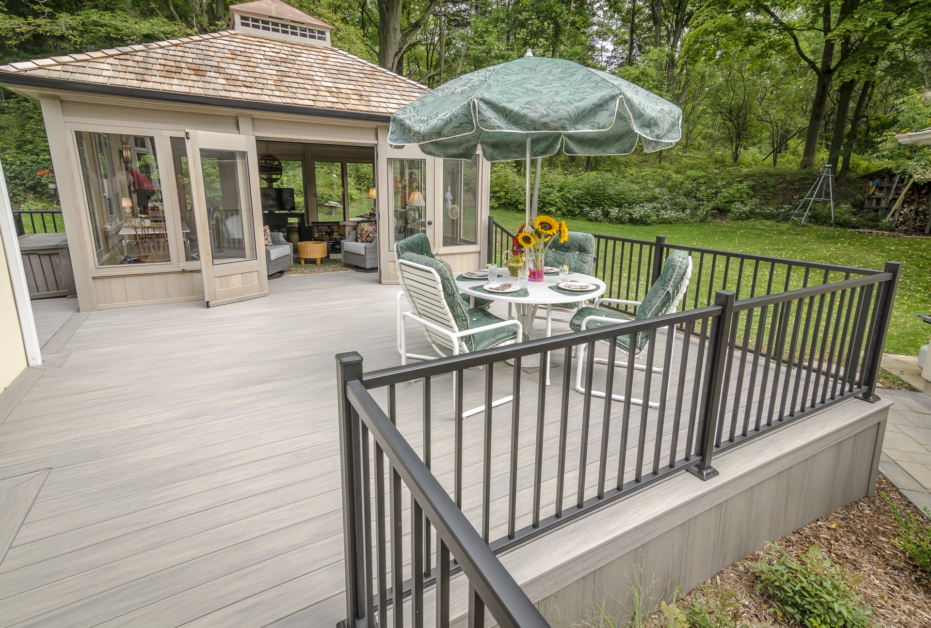 A wooden patio features a sunroom, table with chairs, and umbrella, overlooking a green backyard with trees. Sunflowers add a cheerful touch.