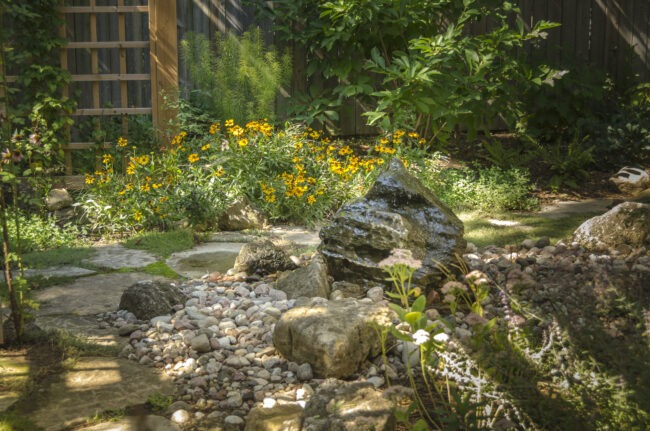 A tranquil garden with lush greenery, blooming yellow flowers, stone path, and rock arrangements create a serene natural landscape.