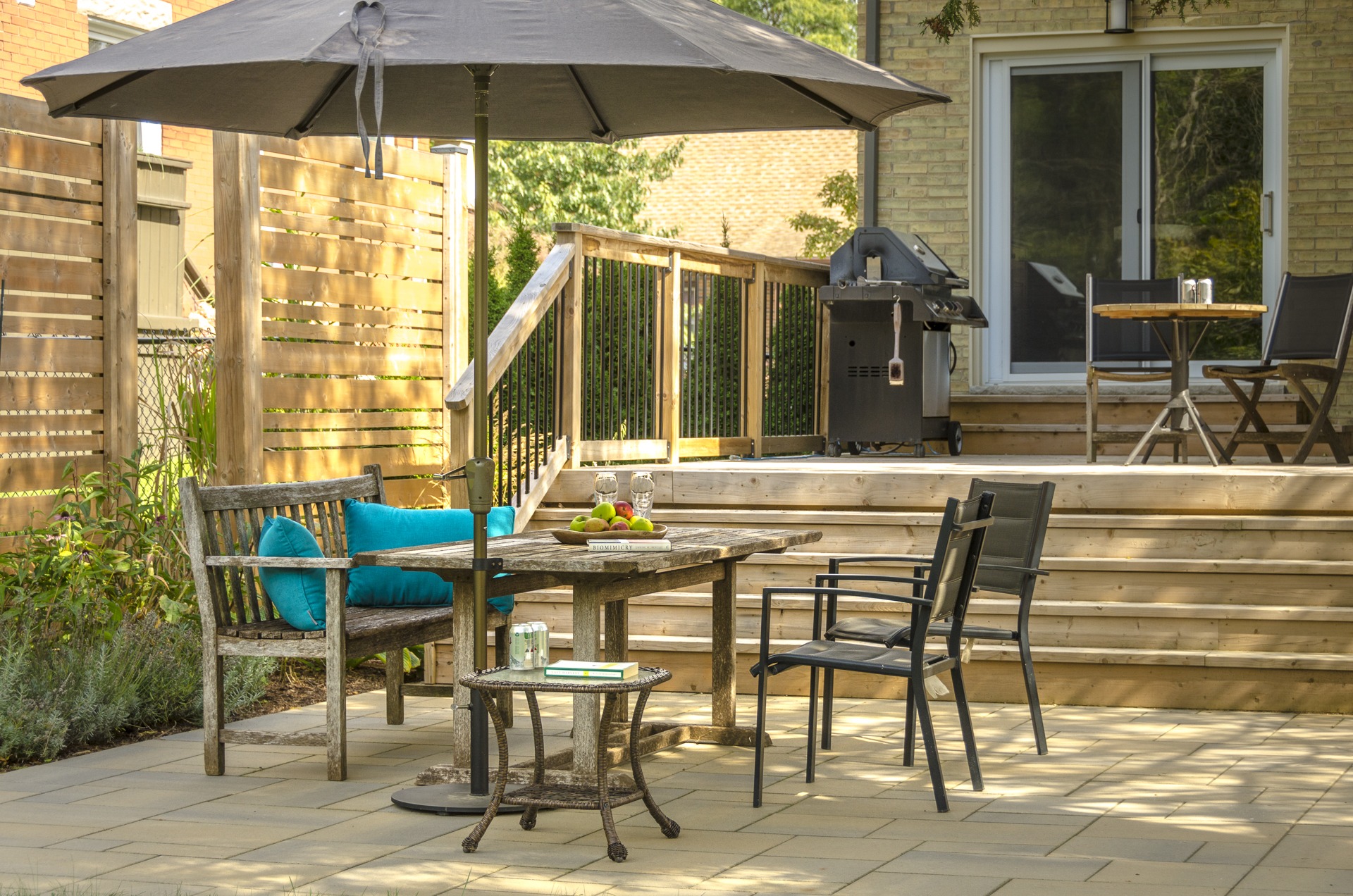 A cozy backyard patio with wooden furniture, blue cushions, a parasol, and a grill. Steps lead to a sliding glass door.