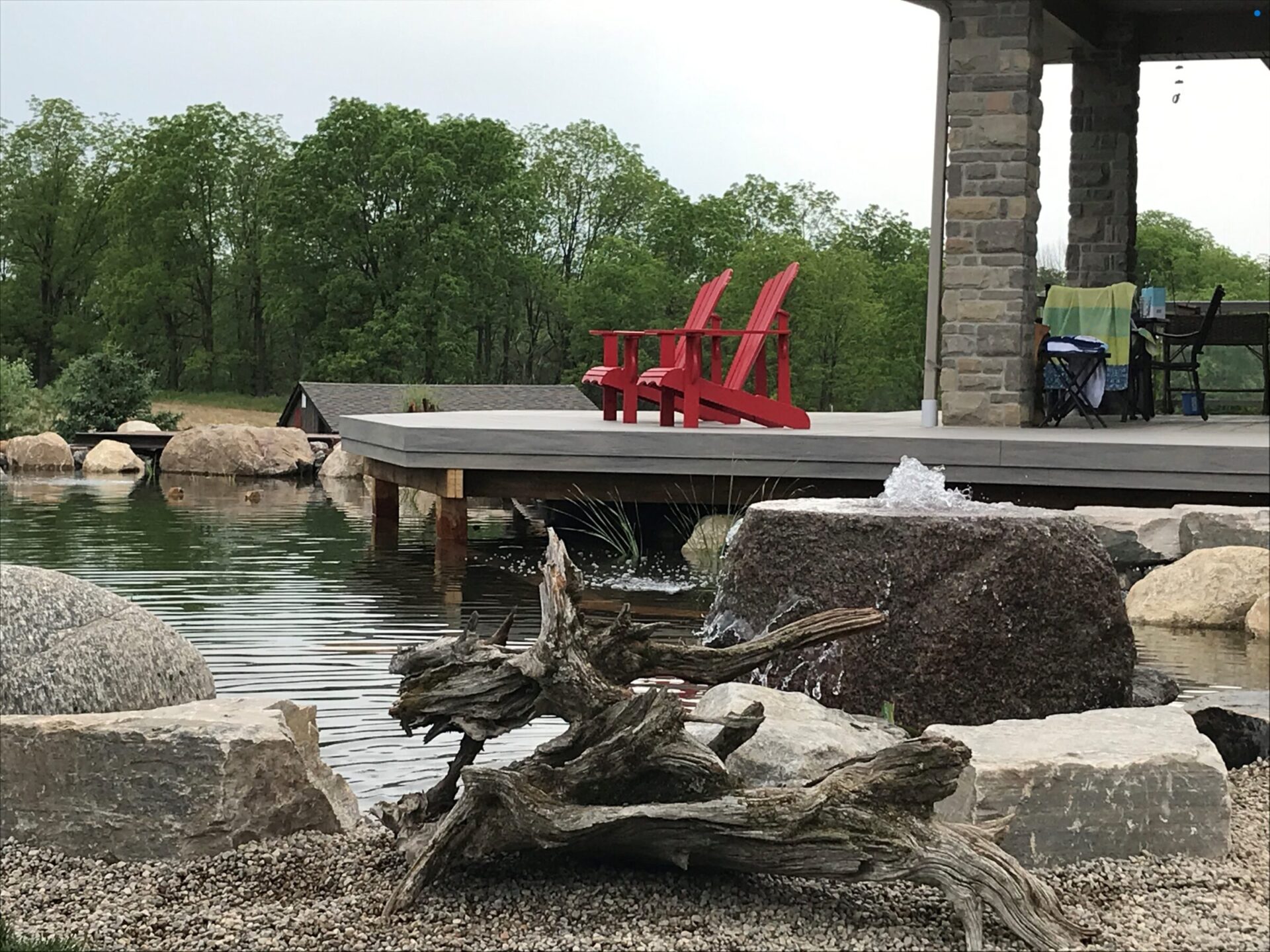 A serene pond with boulders, wooden deck, and red chairs. Trees in the background create a peaceful, natural setting.