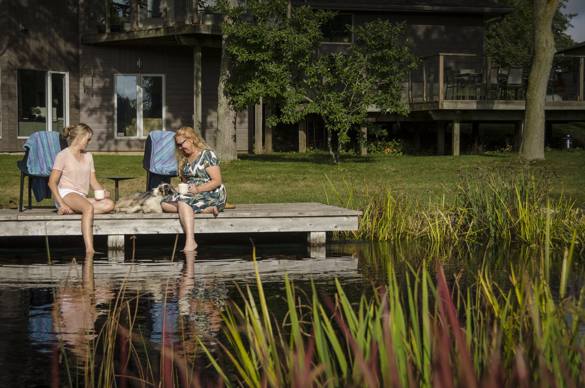 Two people sitting on a dock by a pond, enjoying coffee with a dog, near a wooden house in a green setting.