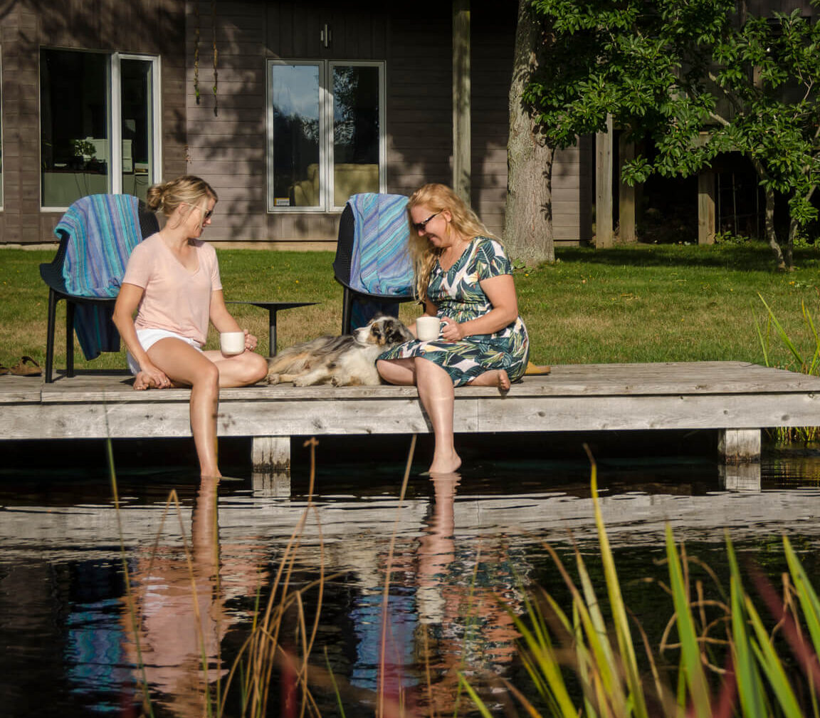 Two people sitting on a dock with a dog, beside a house surrounded by greenery, enjoying a sunny day by the water.