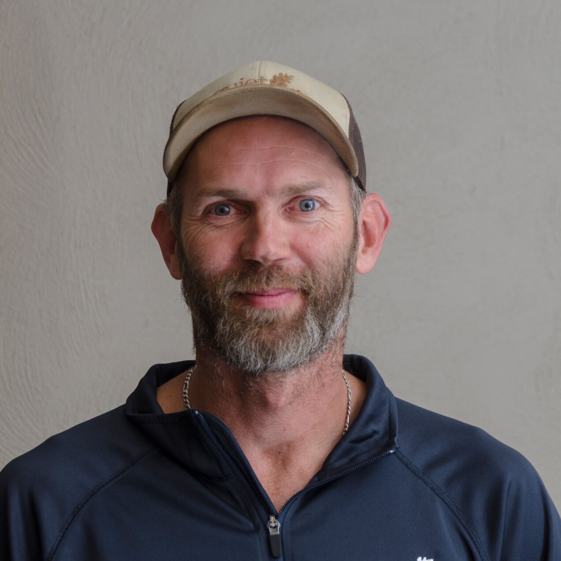 A person with a beard, wearing a cap and a dark zip-up jacket, smiles against a plain background.