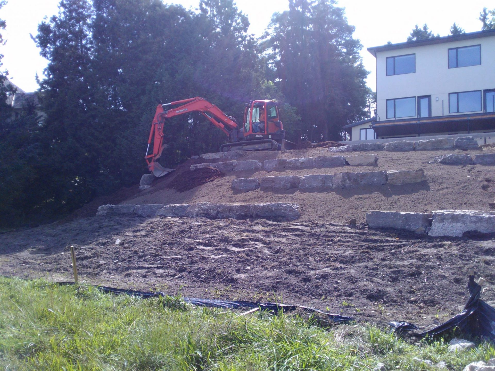 Excavator shaping a steep backyard slope with tiered natural stone retaining walls beside the house