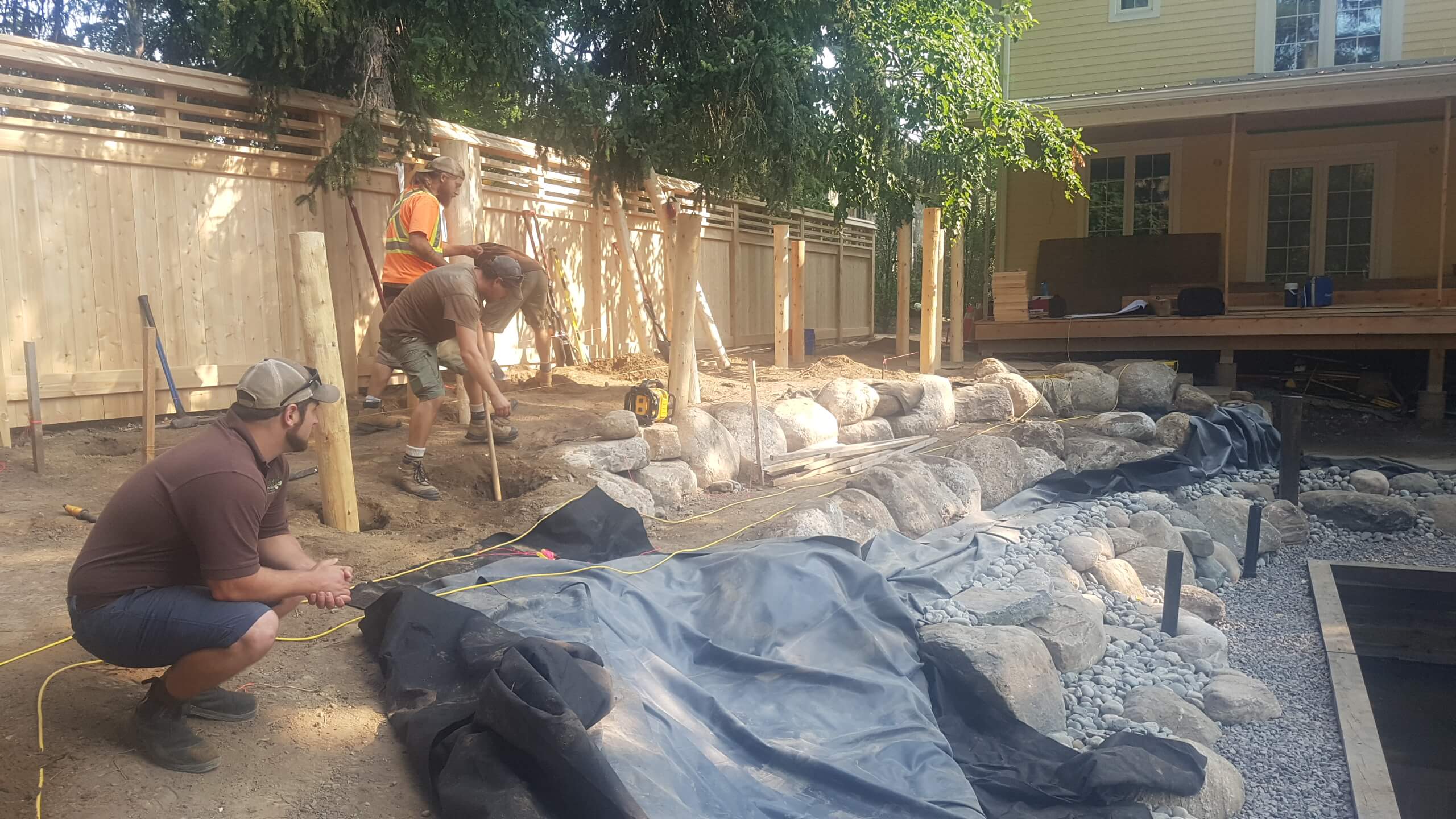 Crew working on the backyard build at The Junction in Guelph, installing posts, stone edging, and pond features beside the rear deck.