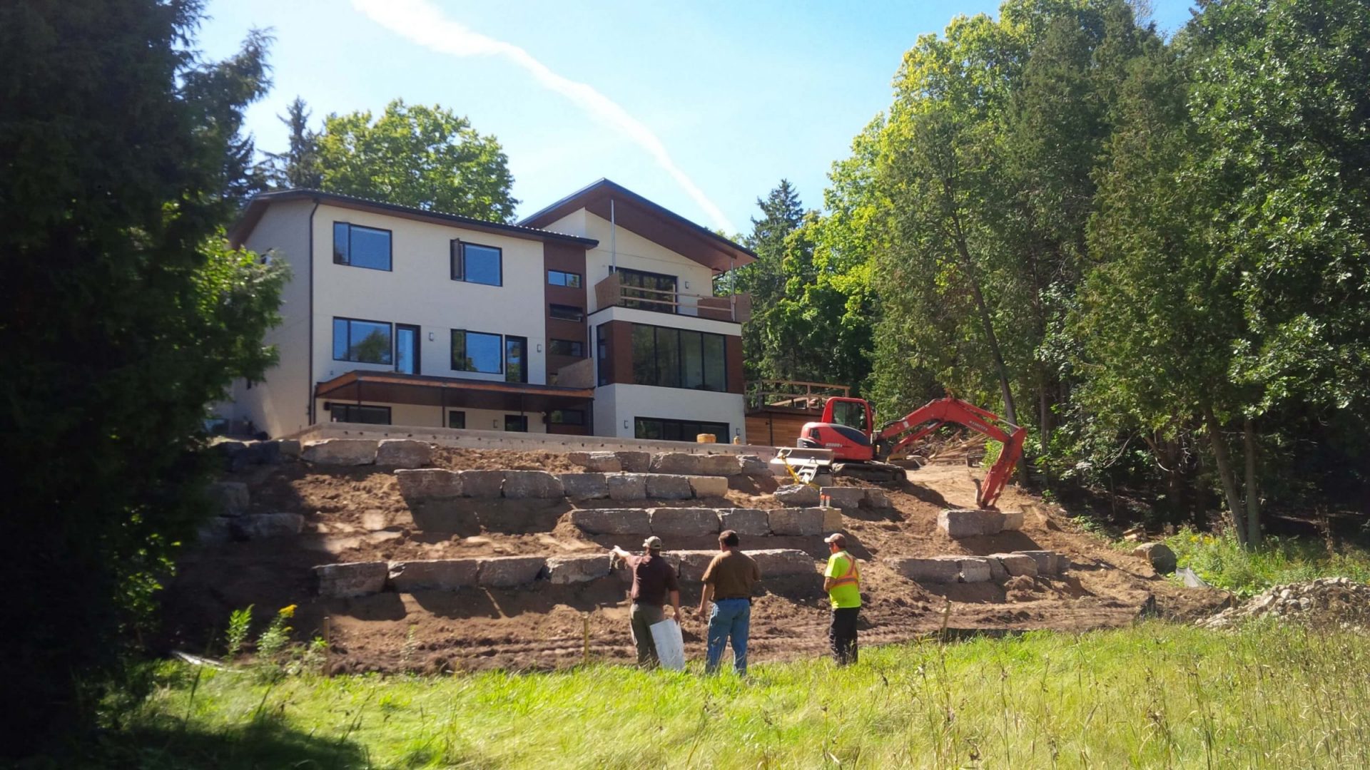 Rear view of the modern home during landscape construction with large natural stone retaining walls, excavation work, and crew members on the sloped backyard