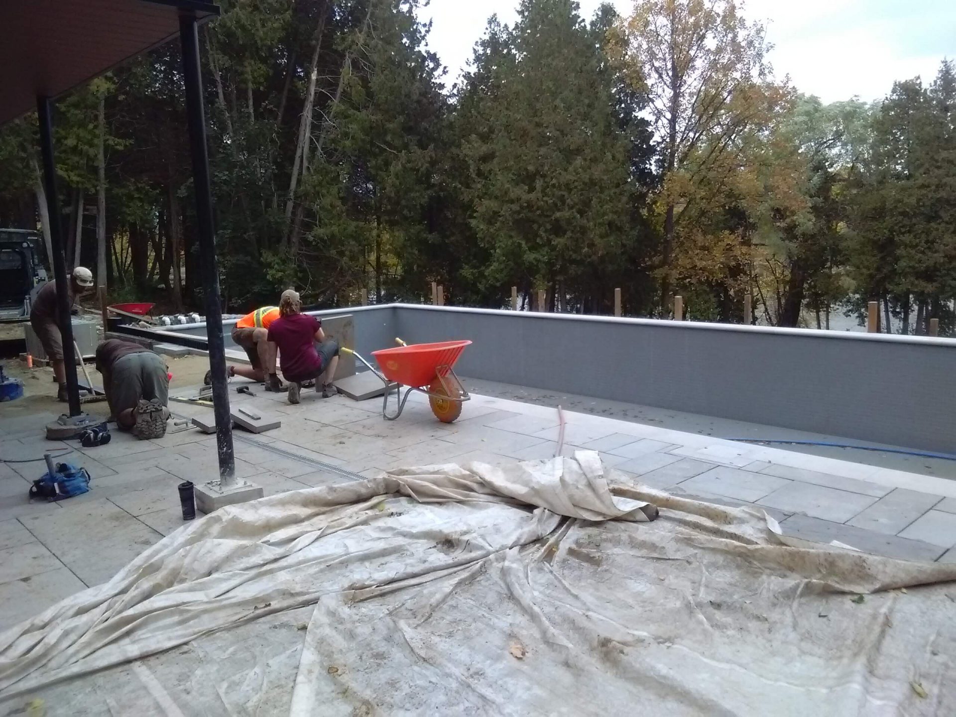 Poolside patio under construction with workers laying stone pavers and a wooded river backdrop beyond