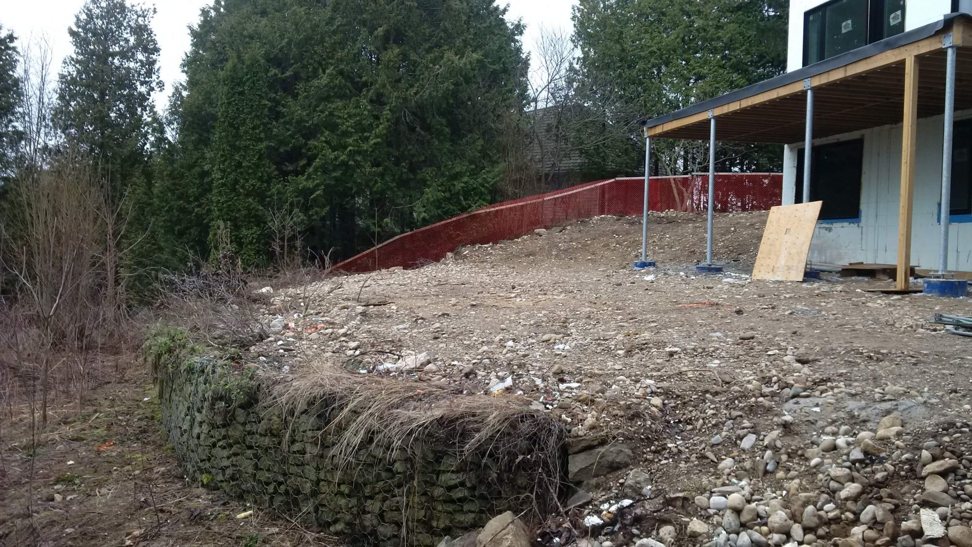 Wide view of the raw sloped backyard with exposed soil, scattered stone, and an existing retaining wall below the house