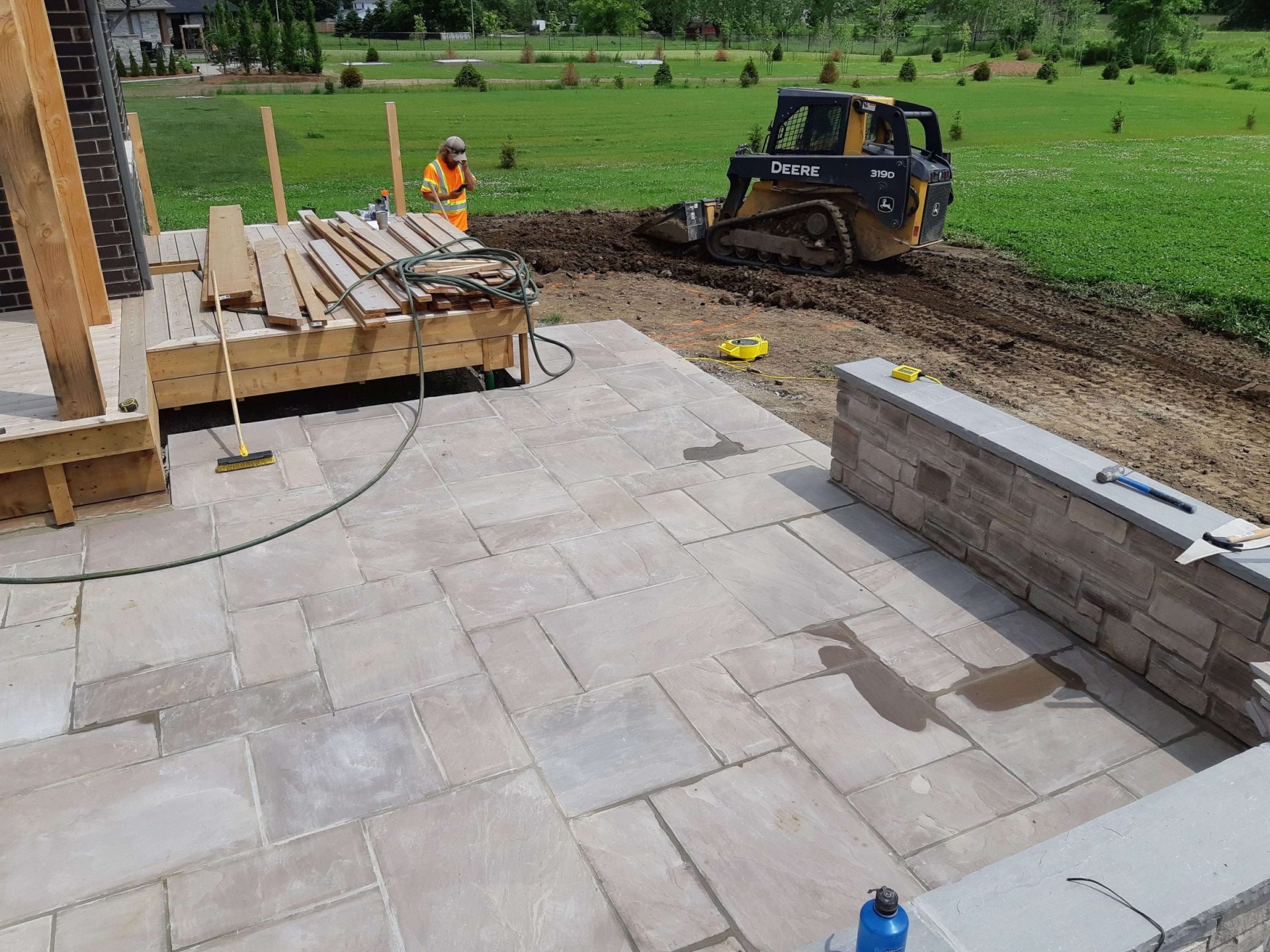 Backyard patio being installed beside the cedar deck with new paving, low wall construction, and grading work underway