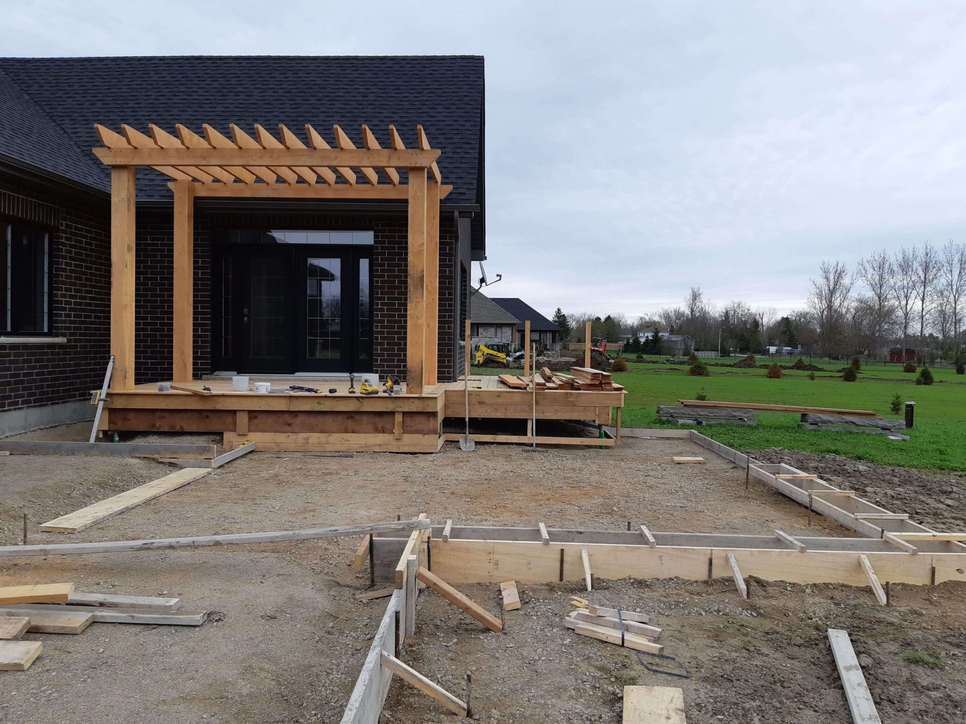 Cedar pergola and rear deck under construction beside the dark brick house with forming in place for the future backyard patio