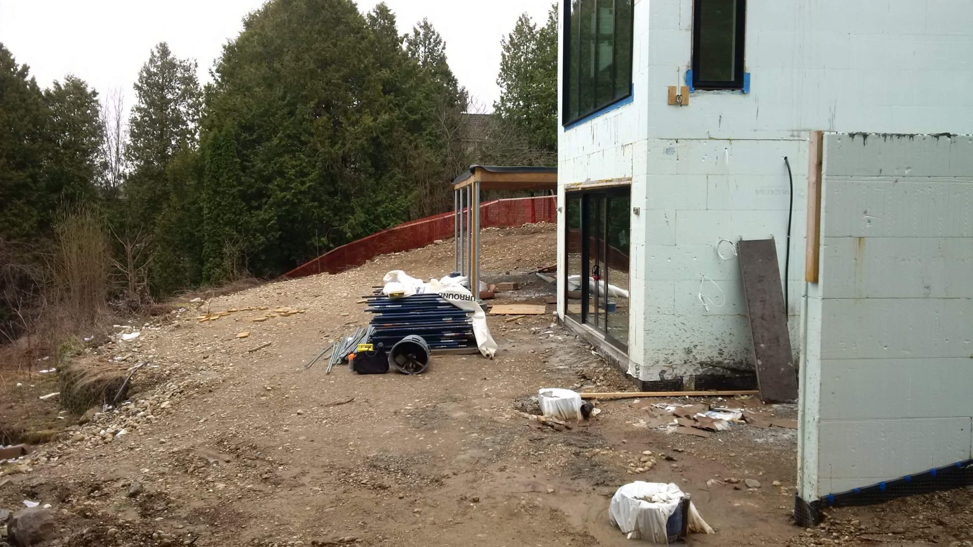 Side view of the house showing a rough sloped backyard with bare soil, construction materials, and a tree-lined edge