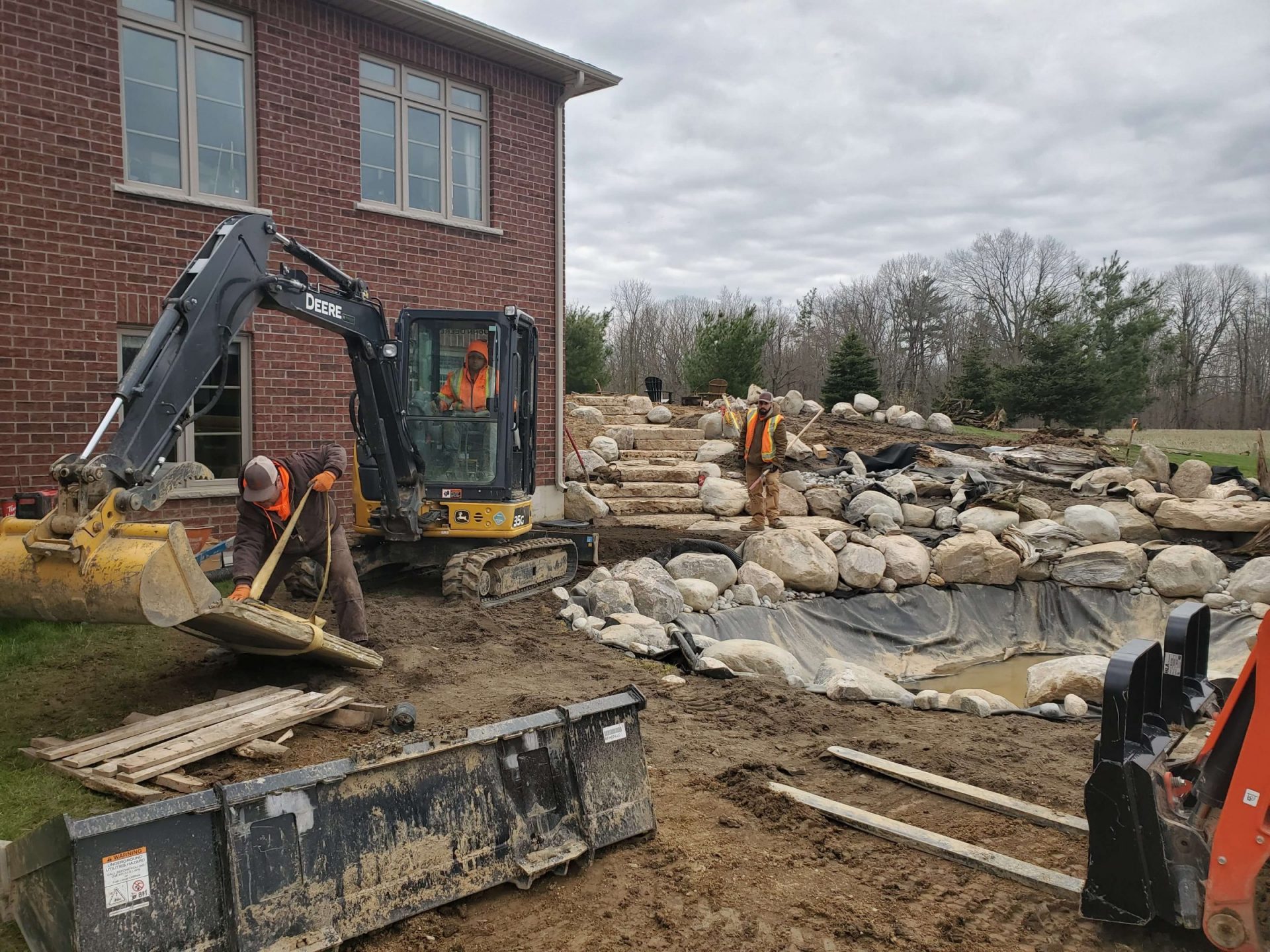 Backyard under construction with an excavator, large boulders, stone steps, and the early shape of the natural pond beside the brick house