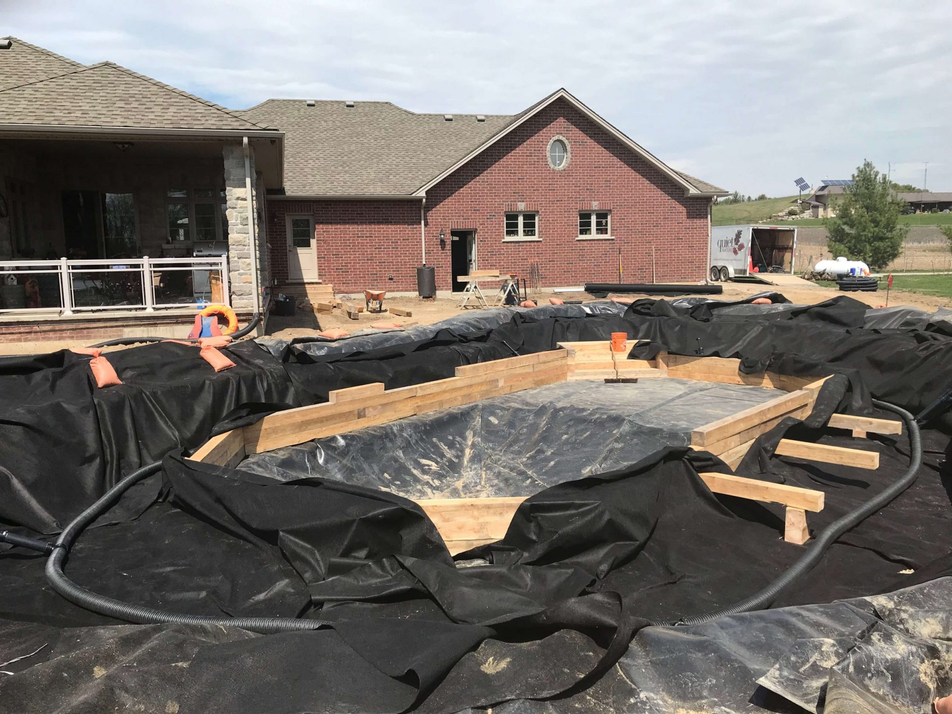 Natural swim pond under construction with black liner, timber framing, and the brick house in the background