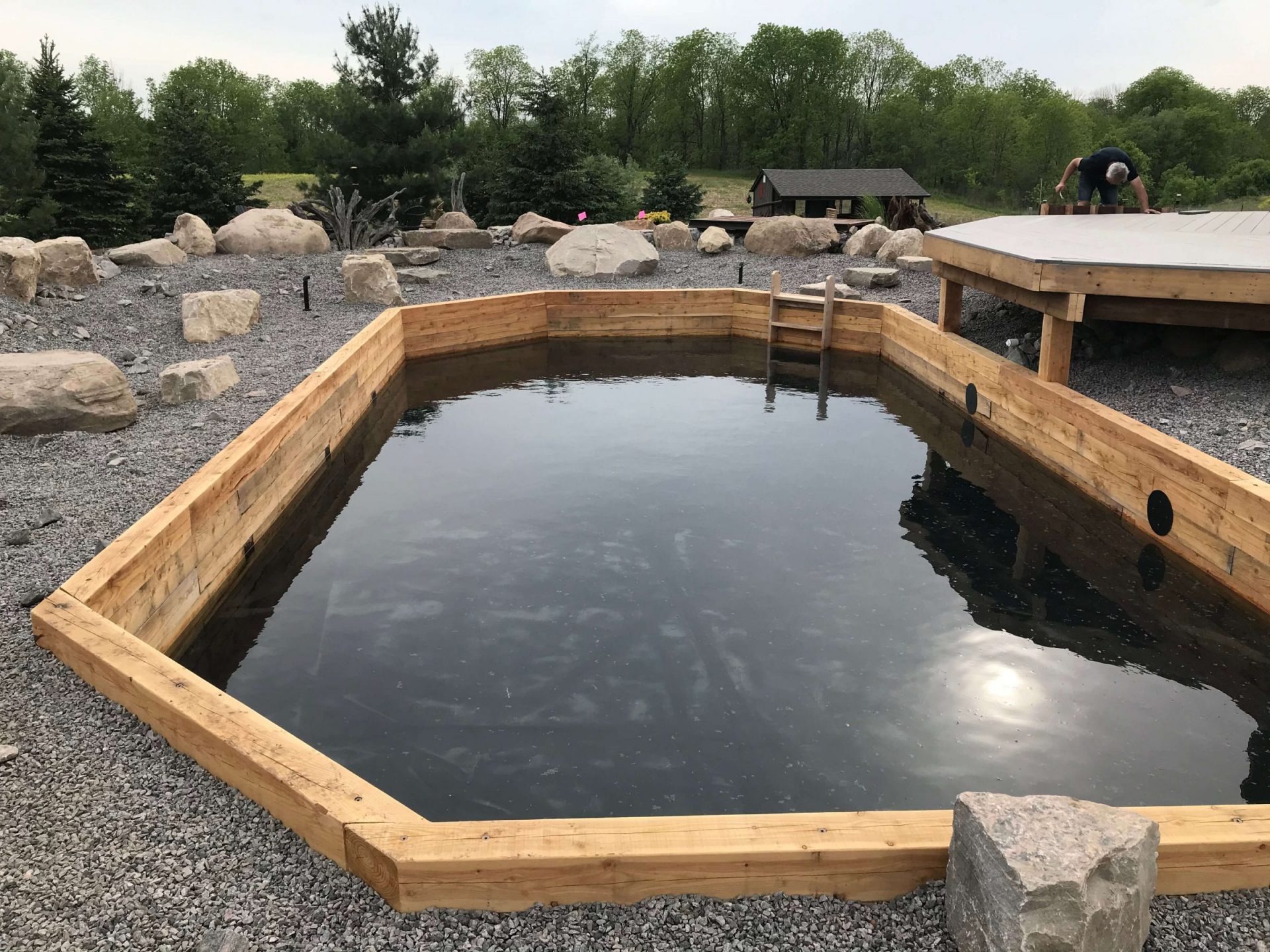 Timber-walled swim pond during construction with gravel surroundings, boulders, and a deck structure beside the water