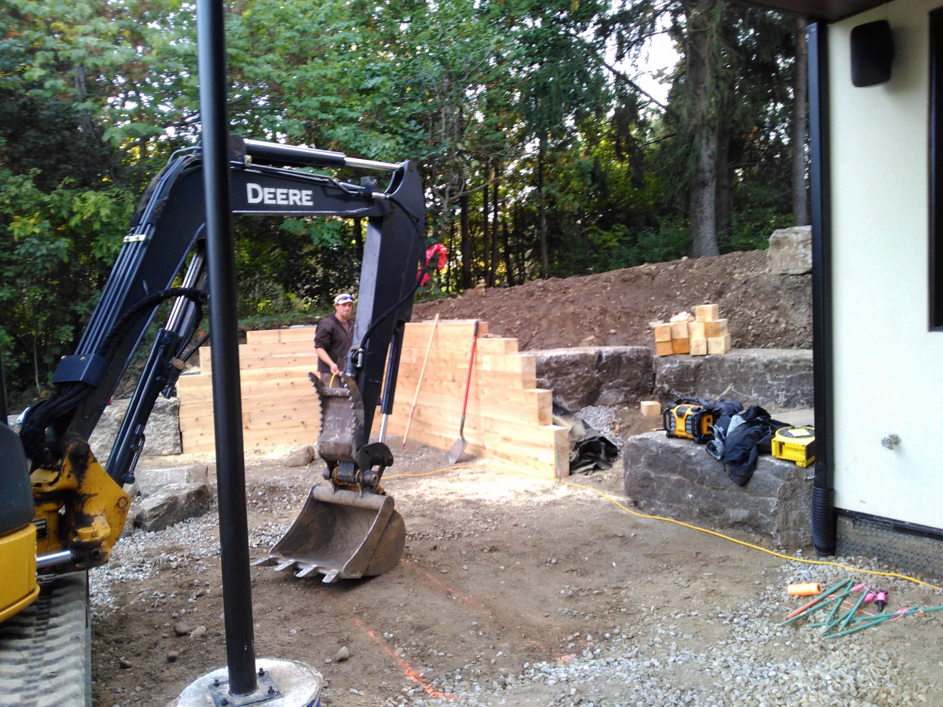 Worker and excavator building a wood stair or retaining structure between stone terraces on the sloped backyard