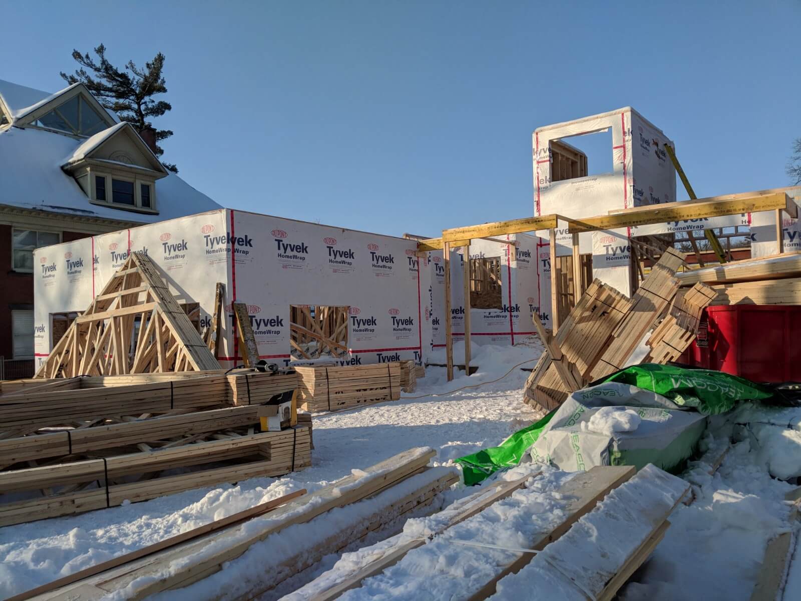 Front area of the house during construction with exposed framing, snow-covered ground, and stacked lumber on site