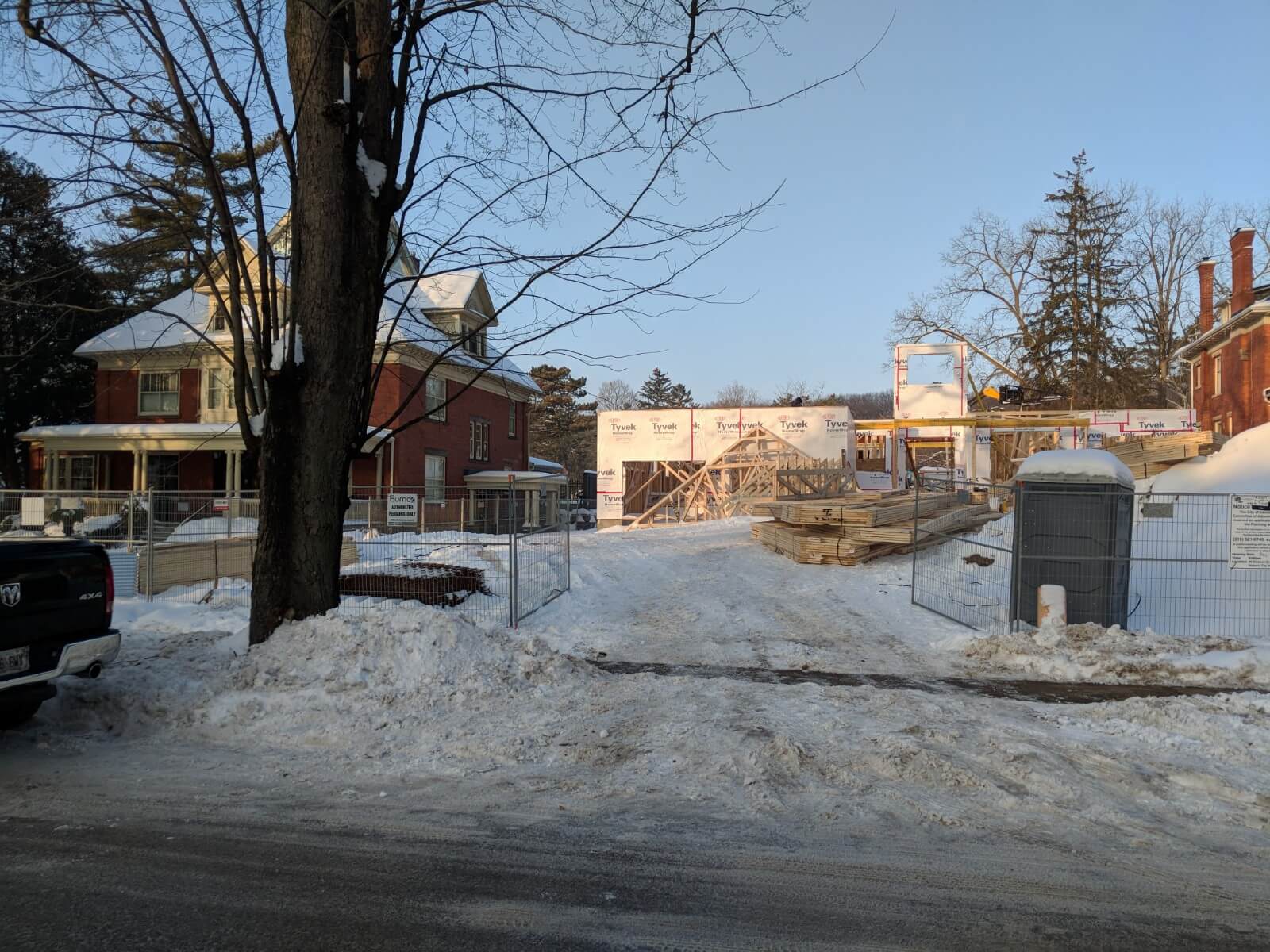 Snow-covered front view of a new home under construction with fencing, building materials, and neighbouring heritage houses