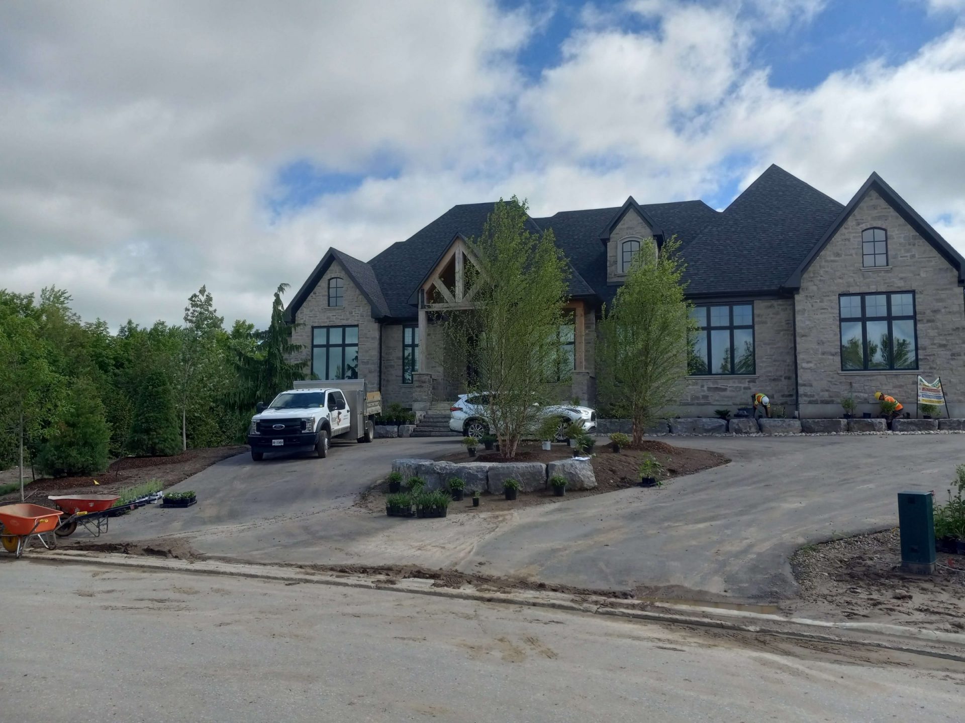 Front view of a large stone home with unfinished driveway edges, rough front landscaping, and early planting work in progress