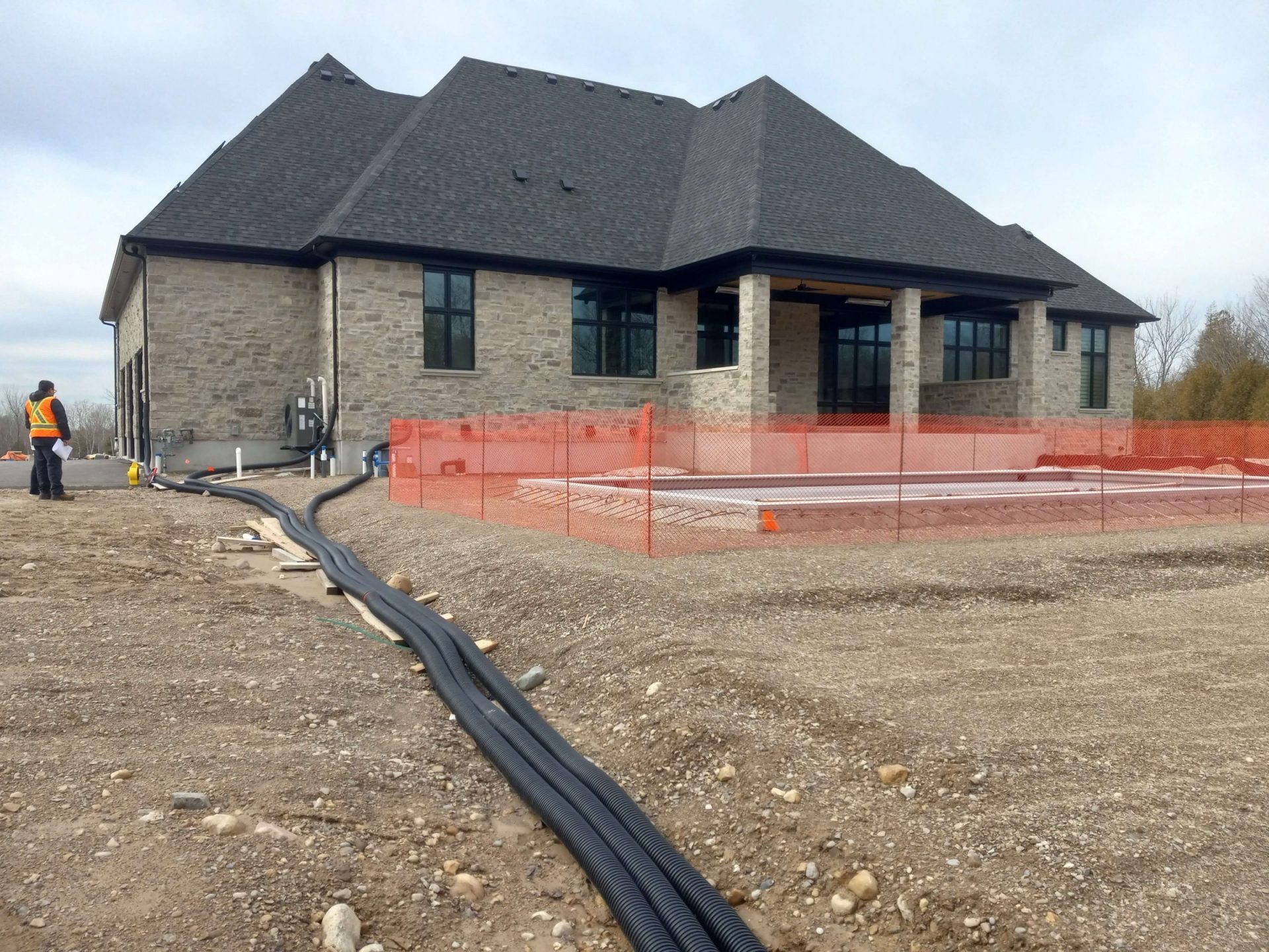 Rear of the house during construction showing pool installation, temporary safety fencing, and exposed drainage lines running through the rough-graded yard