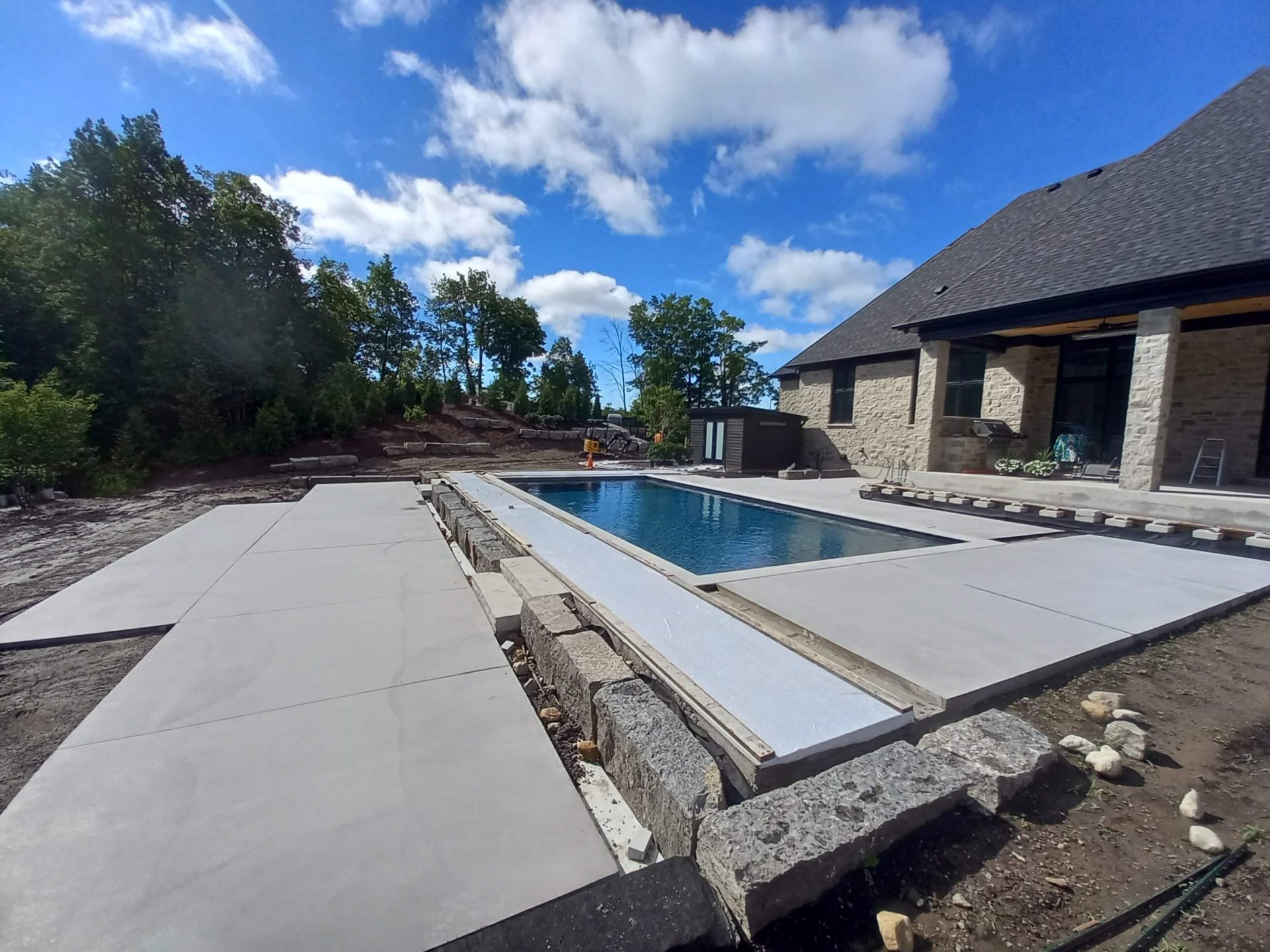 Pool and concrete patio under construction with armour stone retaining, unfinished edges, and backyard grading still underway