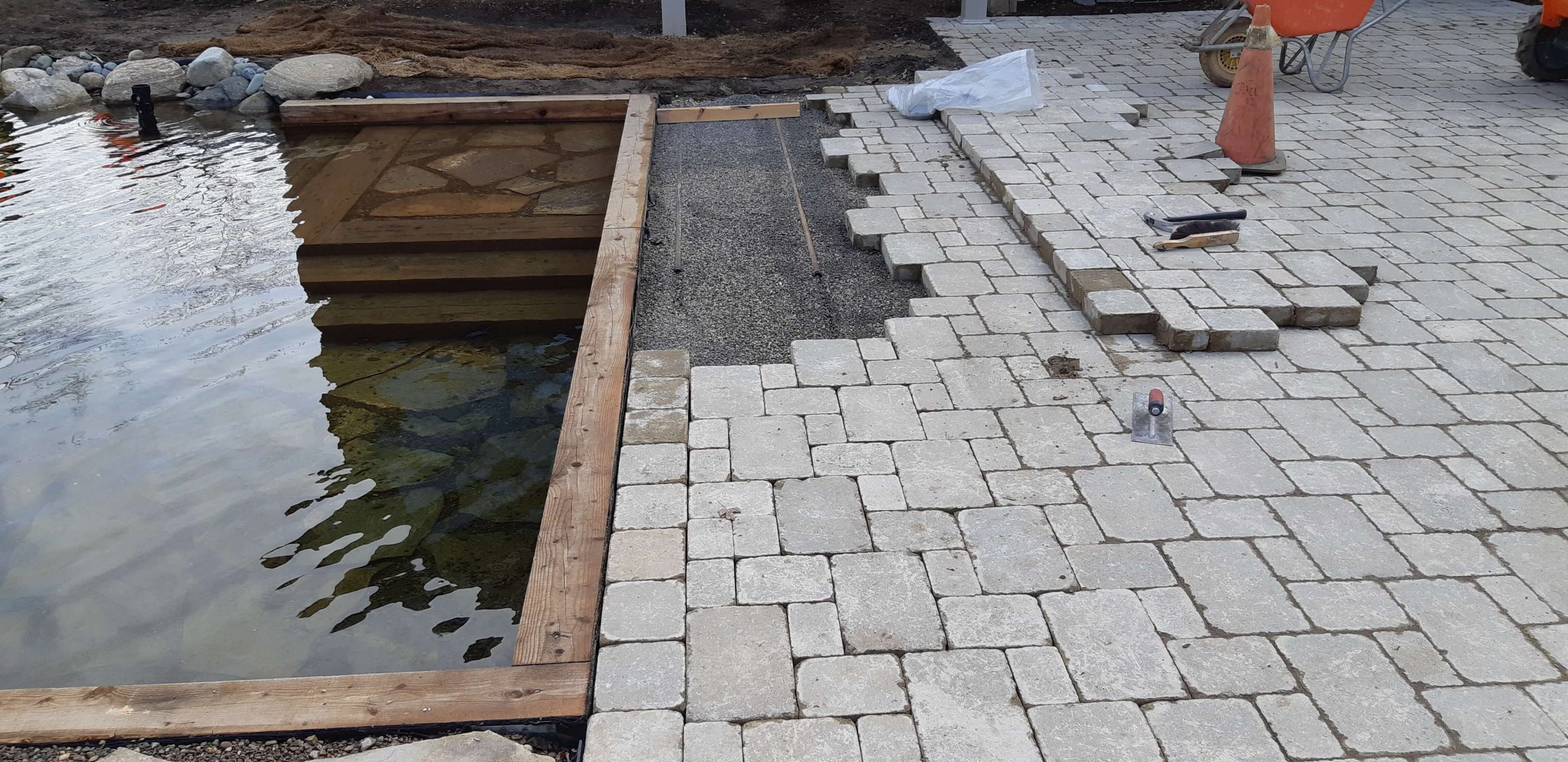 Close view of the natural swim pond edge during construction with timber walls, underwater stone steps, and paver patio installation beside the water