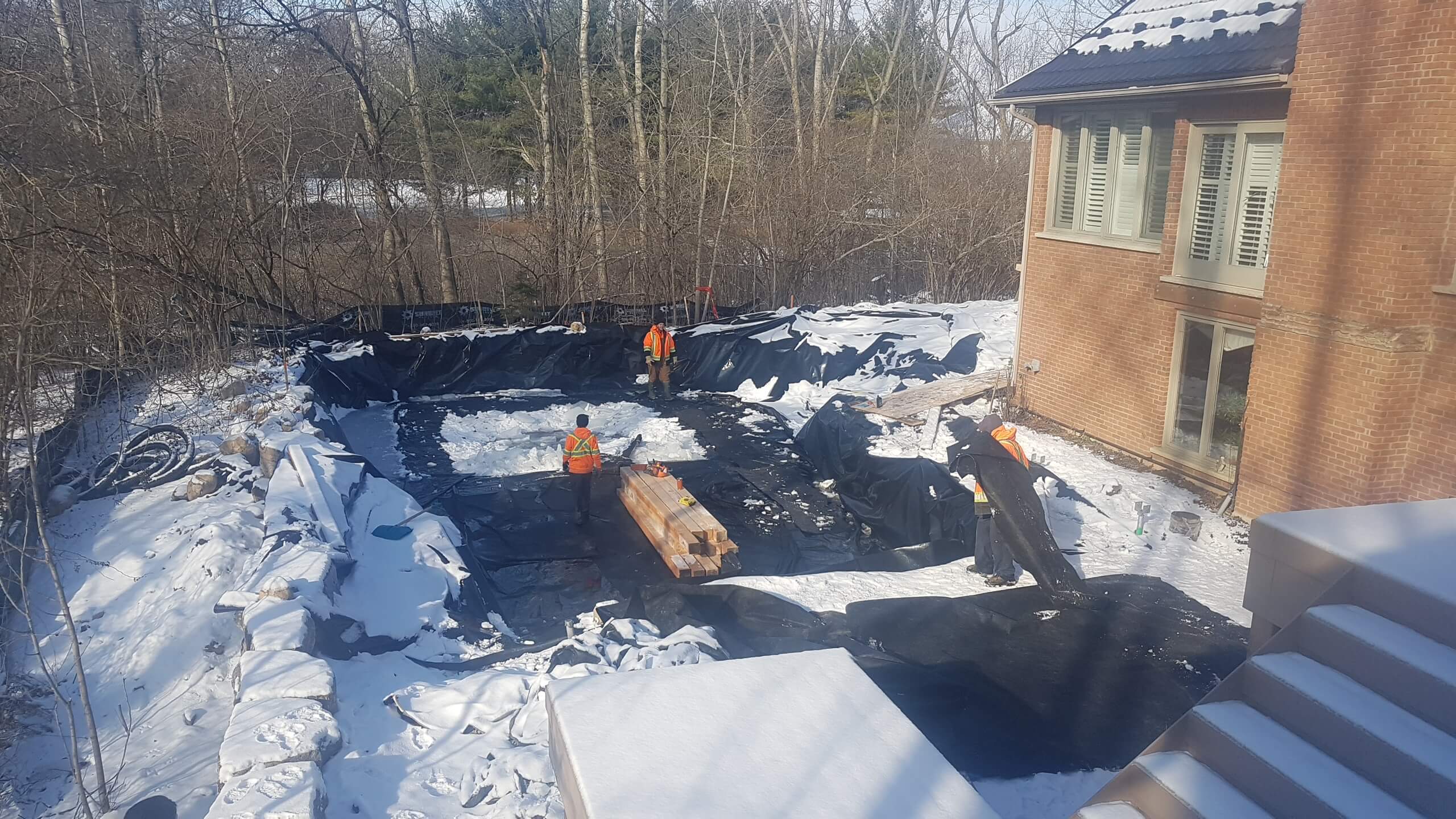 Backyard construction in winter with the natural swim pond excavation lined in black membrane beside the brick house