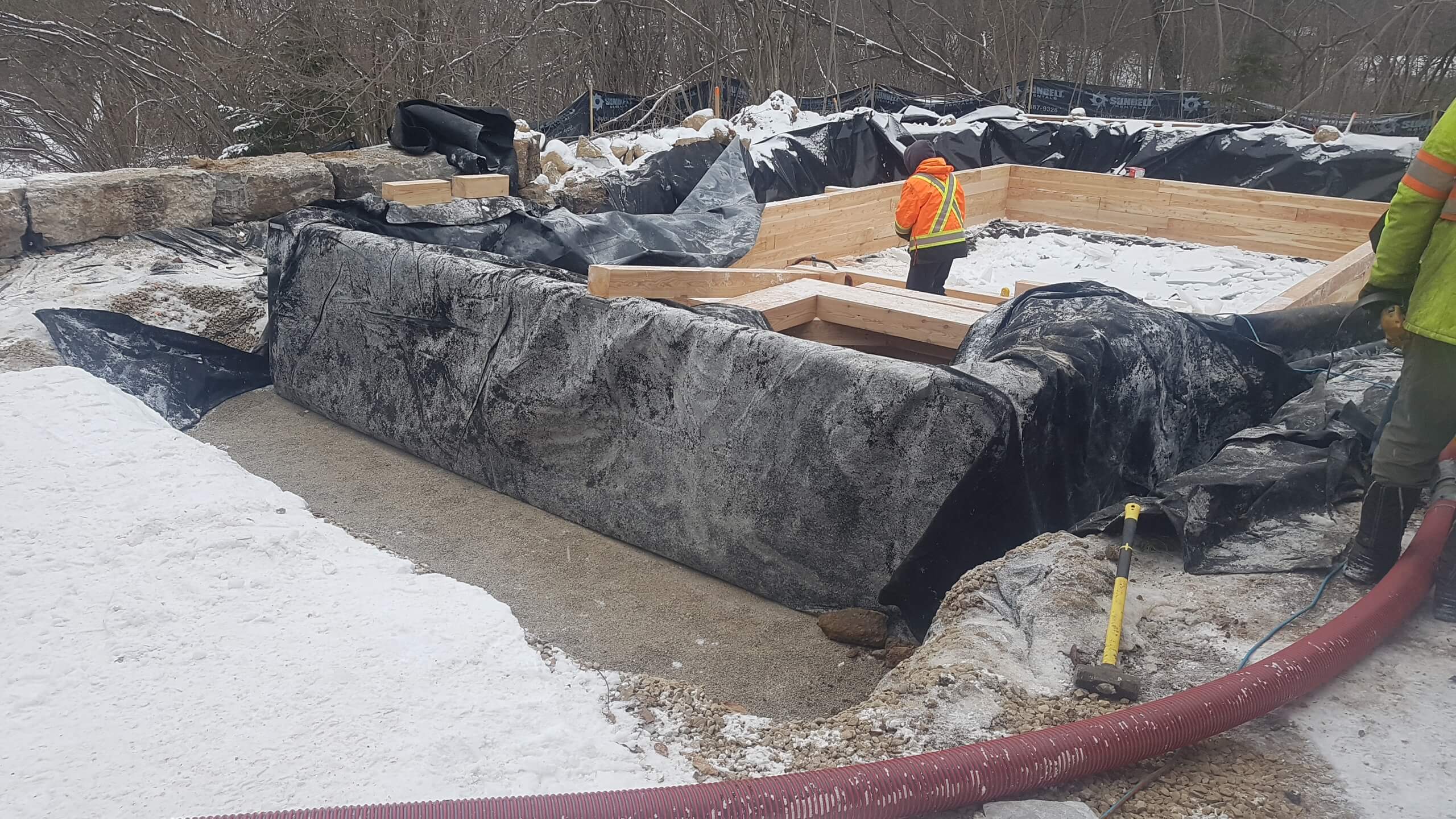 Natural swim pond under construction with timber retaining walls, black liner, and workers shaping the swimming area in winter